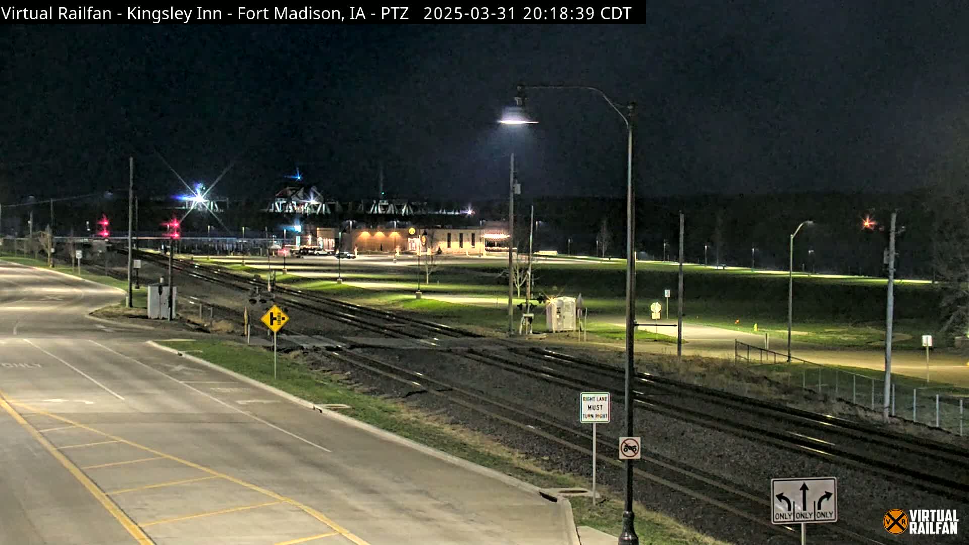A nighttime view of a railroad crossing near a building, with a road intersecting the tracks under clear skies.