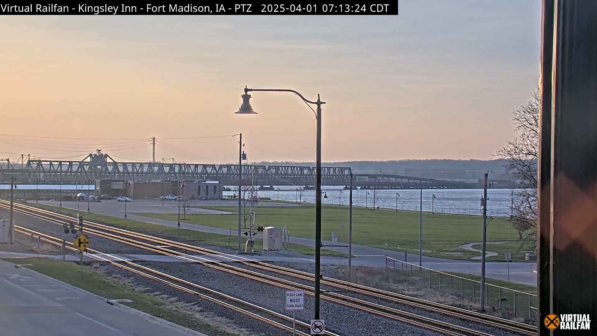 A long metal bridge spans a calm river at sunset, with train tracks and a grassy area in the foreground.