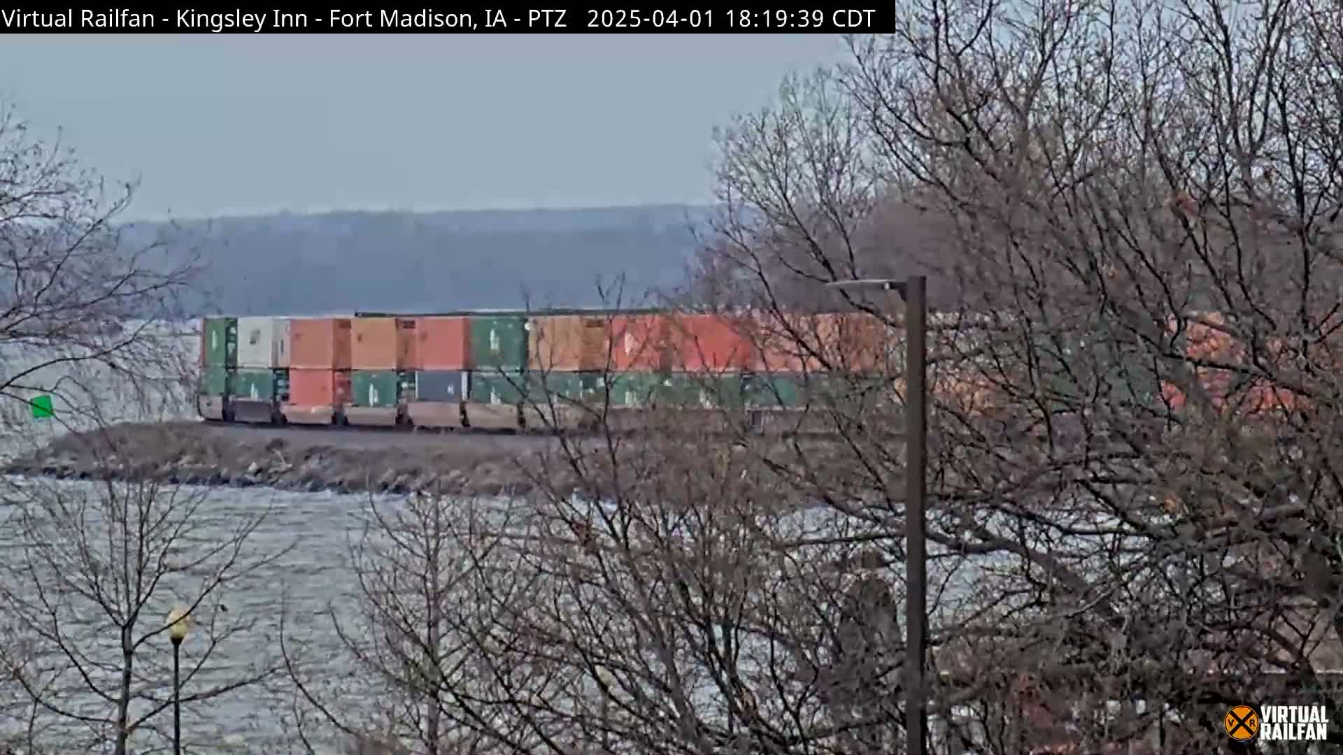 A train carrying a long line of colorful shipping containers sits by a choppy body of water under a cloudy sky, partially obscured by bare, winter trees.