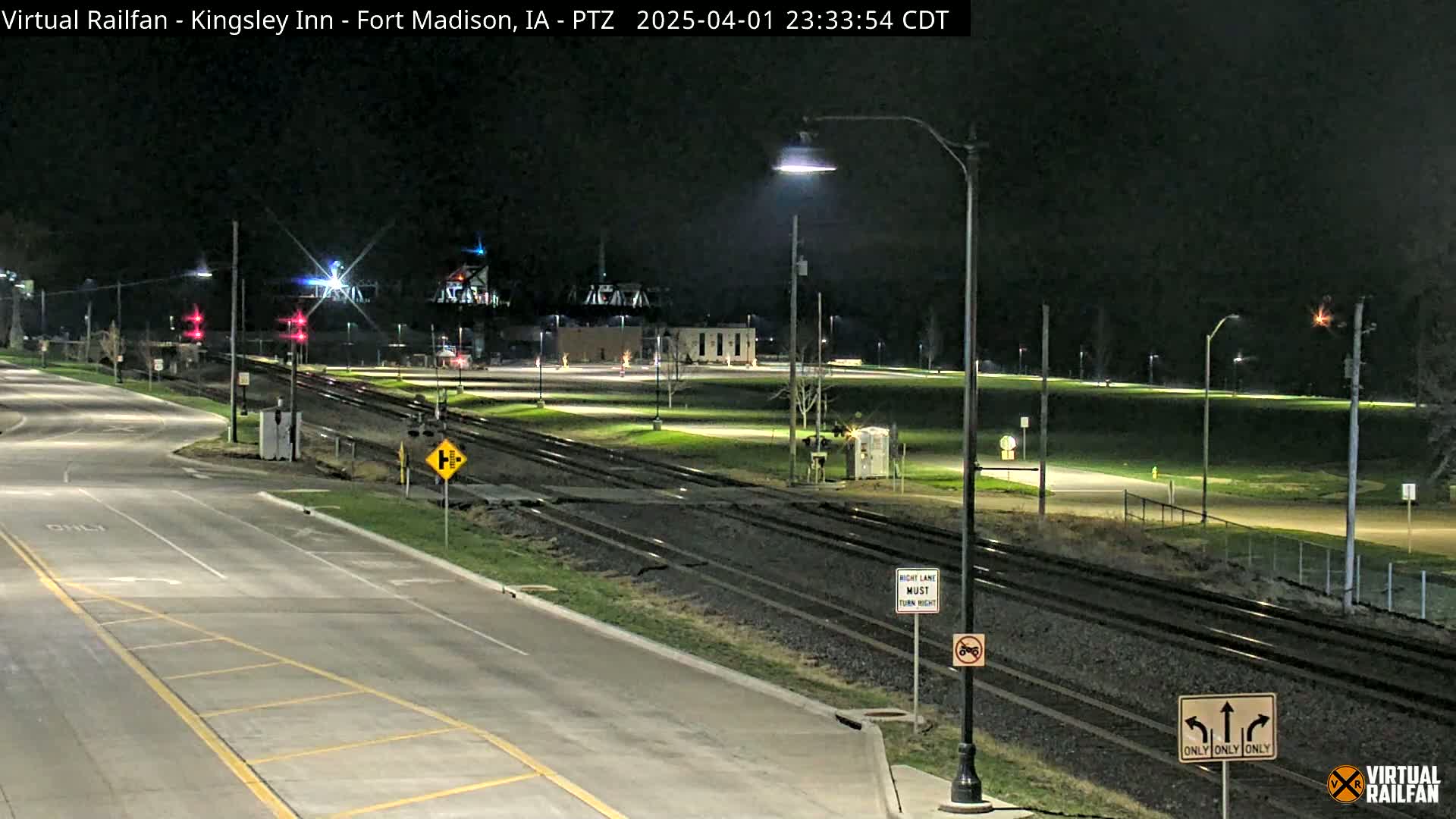 A nighttime view of a paved road intersecting with railroad tracks, under artificial light, with a grassy area beyond.