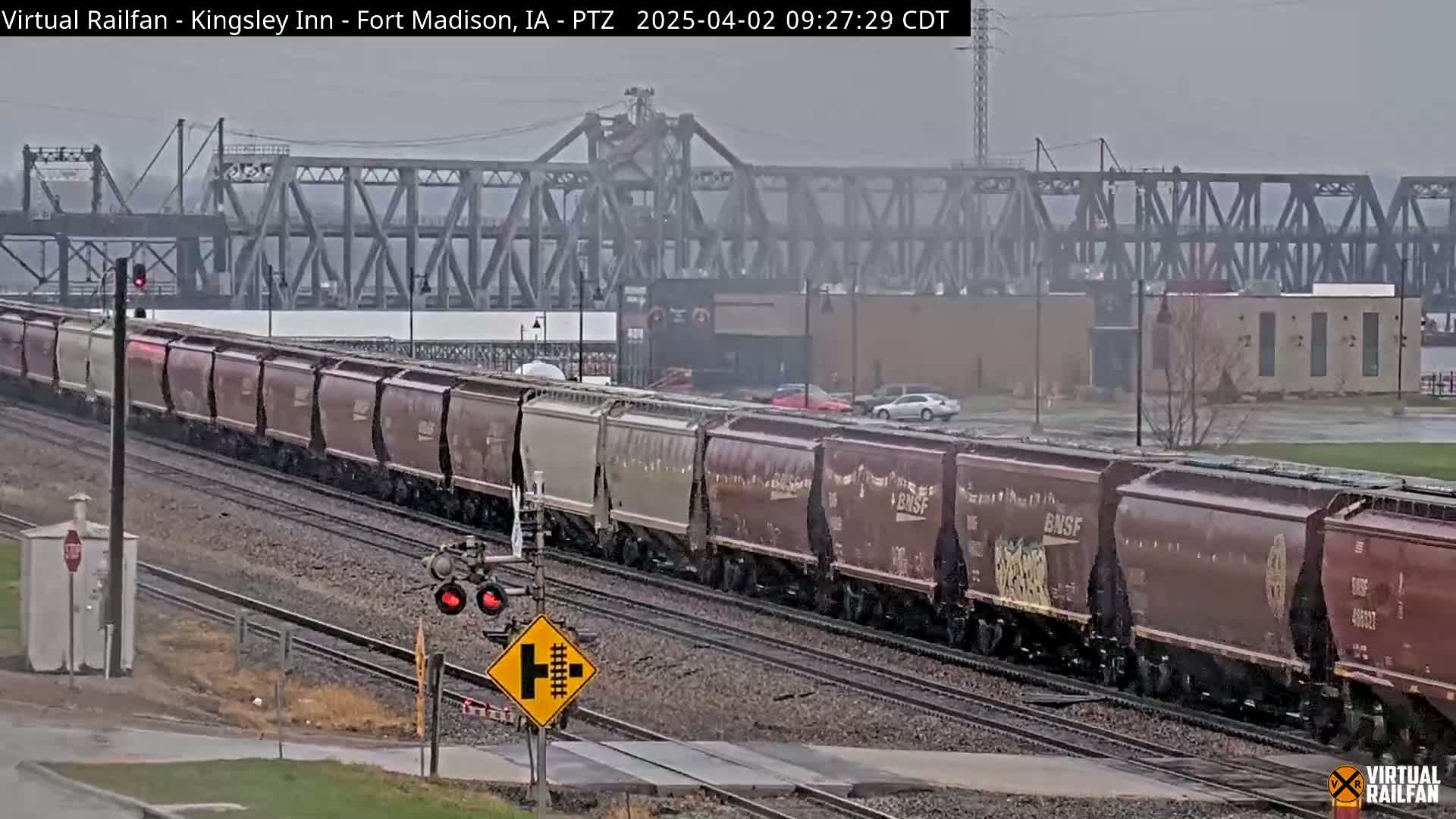 A long freight train passes under a metal bridge on an overcast day.