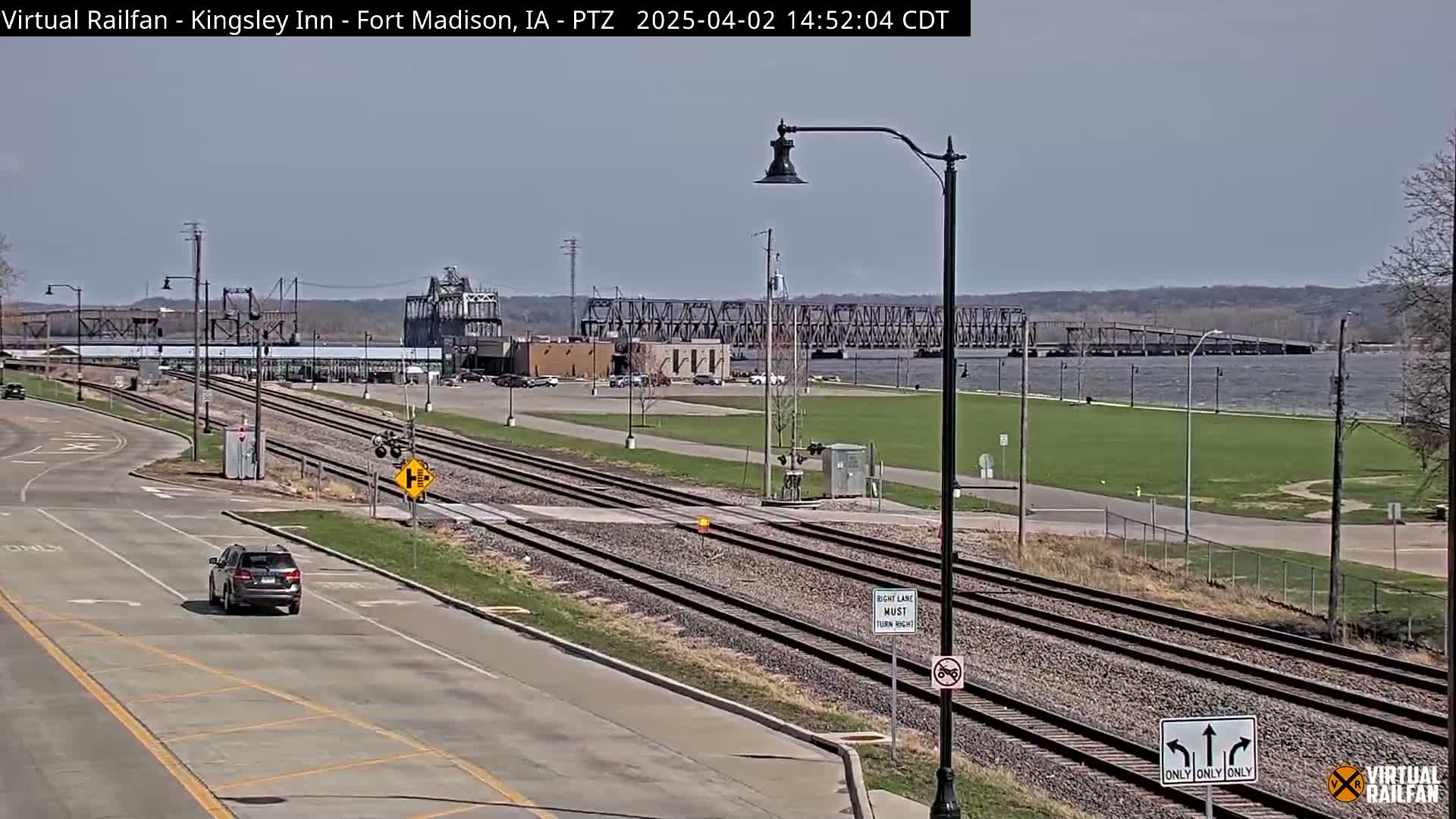 A mostly sunny day reveals a view of train tracks crossing a road near a river, with a bridge visible in the distance, and a single car driving on the road.