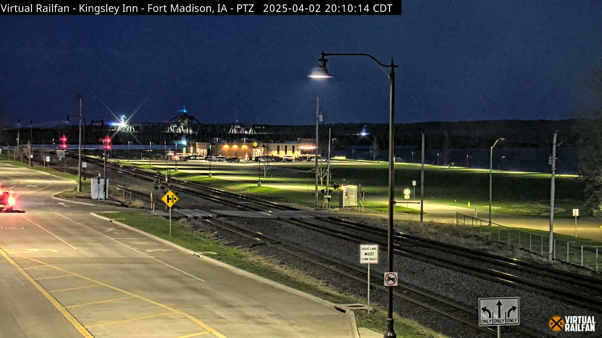 A nighttime view of railroad tracks intersecting a road near a building and a body of water under a dark sky.