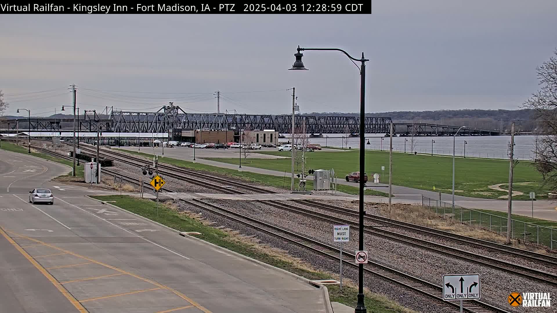 A view of railroad tracks intersecting a road, with a large metal bridge in the background under a cloudy sky.
