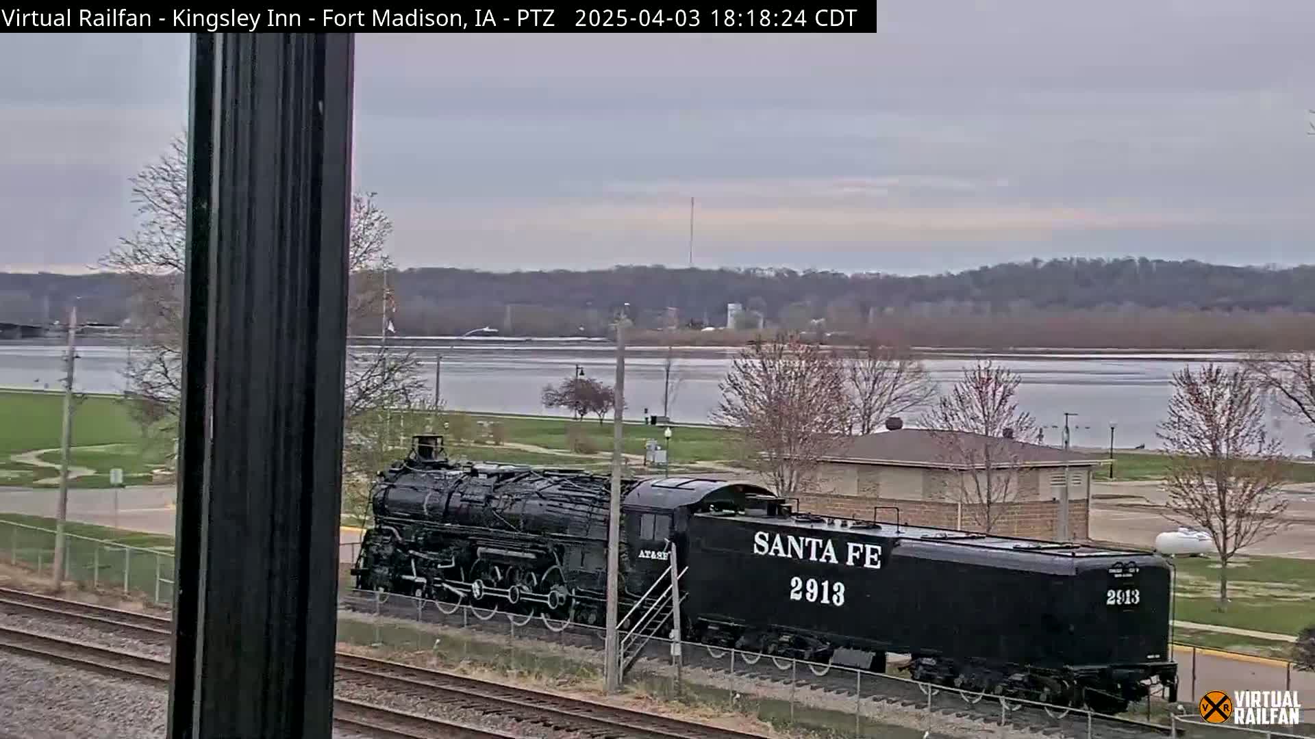 A black Santa Fe 2913 steam locomotive sits on railroad tracks near a river under an overcast sky.