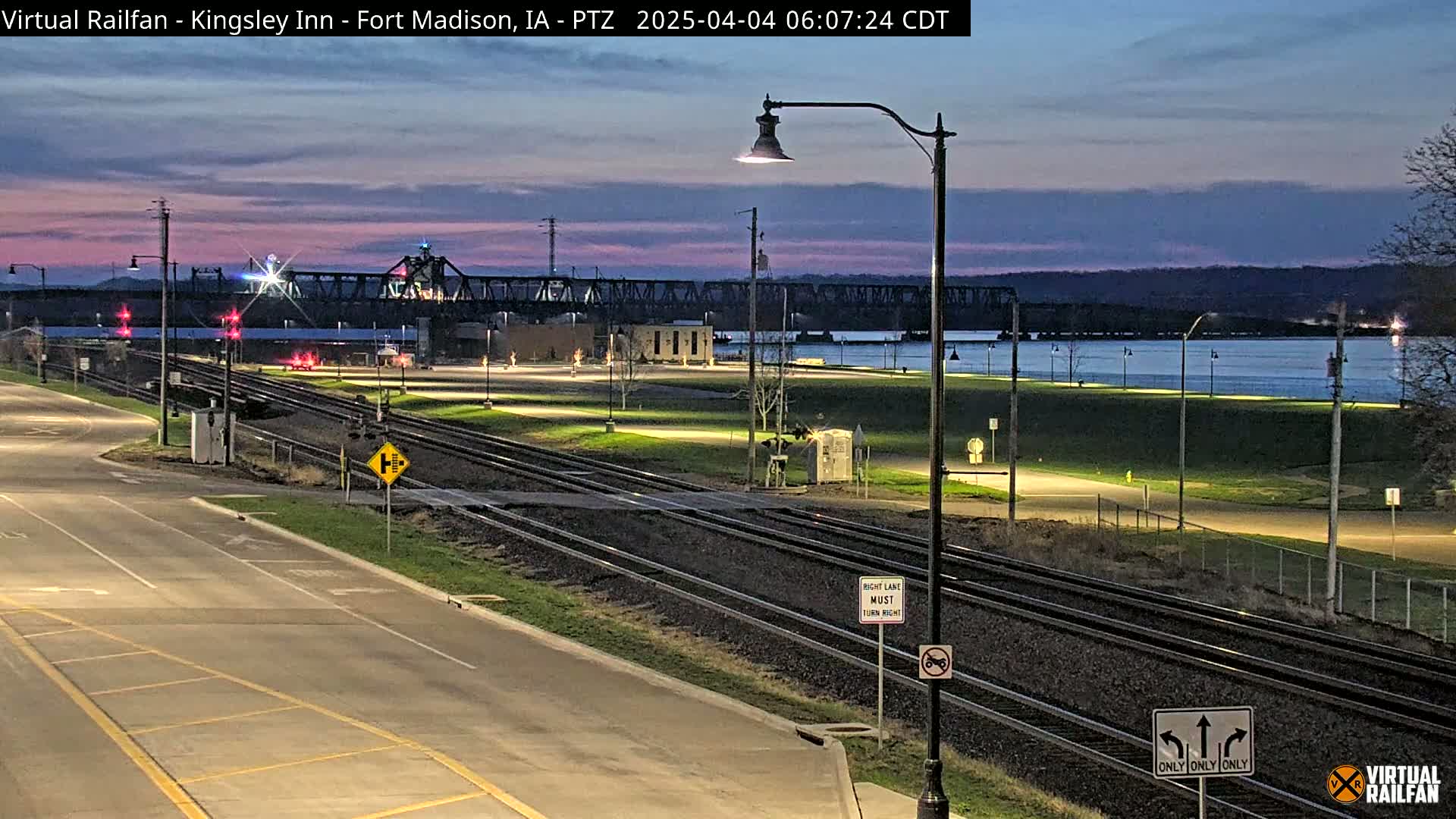 A nighttime view of train tracks intersecting with a road near a river, under a twilight sky.