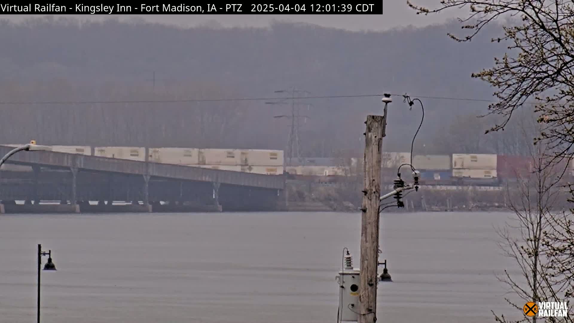 A hazy, overcast day reveals a train on a bridge over a wide body of water, with leafless trees in the foreground.