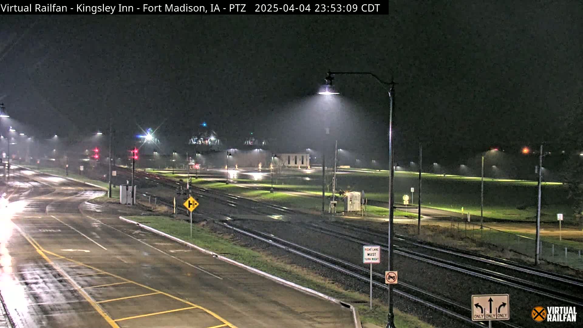 A rain-slicked road and railroad tracks intersect at a nighttime intersection under streetlights.
