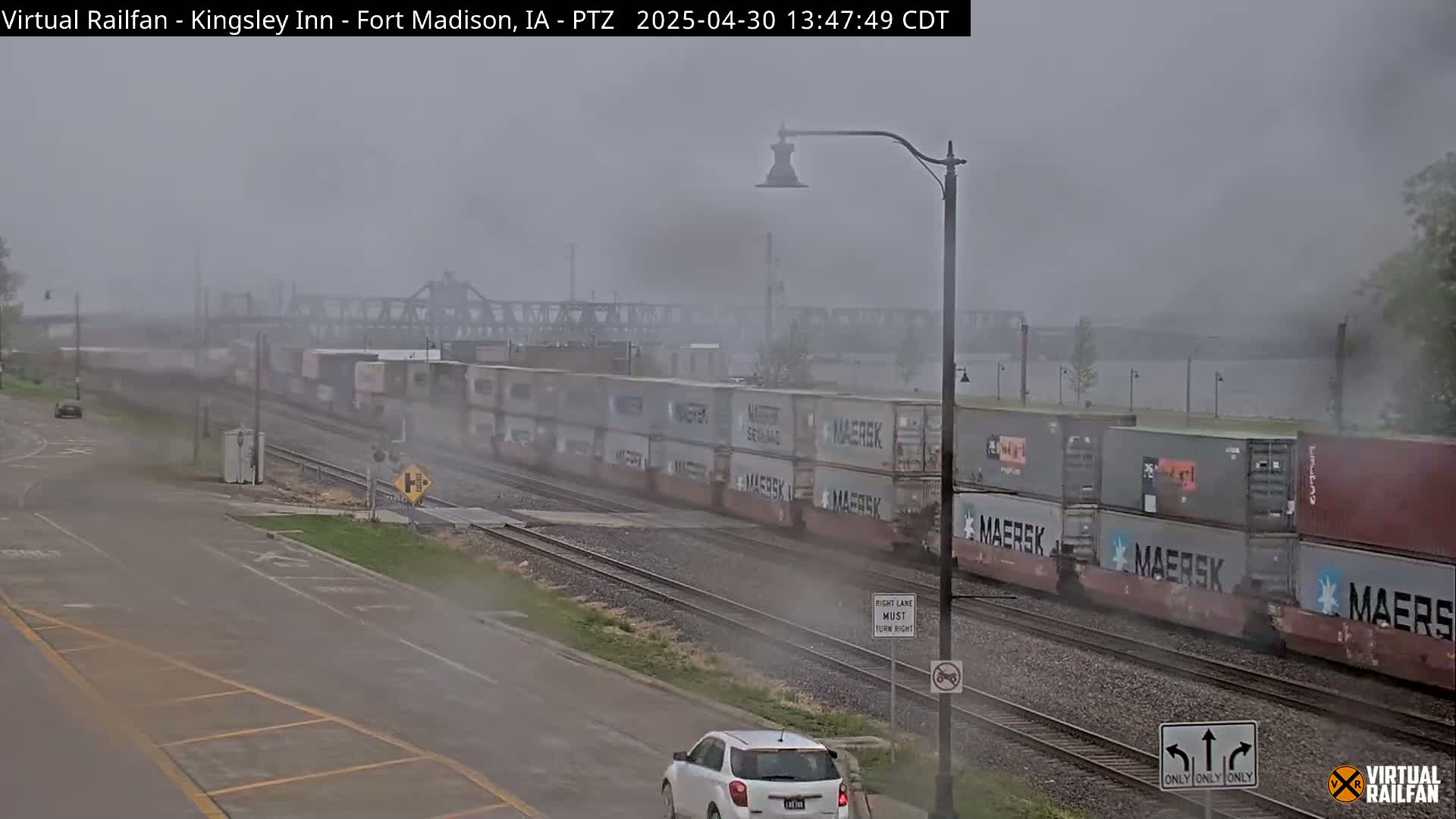 A long freight train carrying numerous shipping containers passes under an overcast sky, alongside a road where a car is visible.