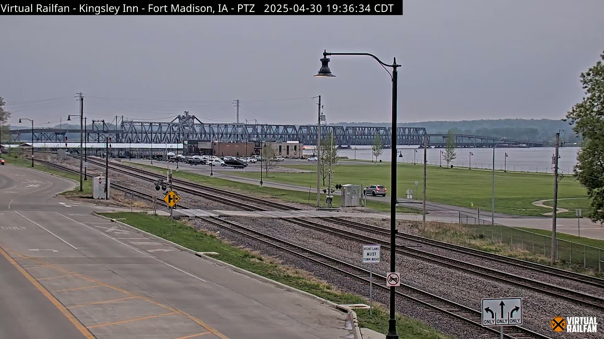 A view of railroad tracks crossing a road near a river, with a large bridge in the background under an overcast sky.