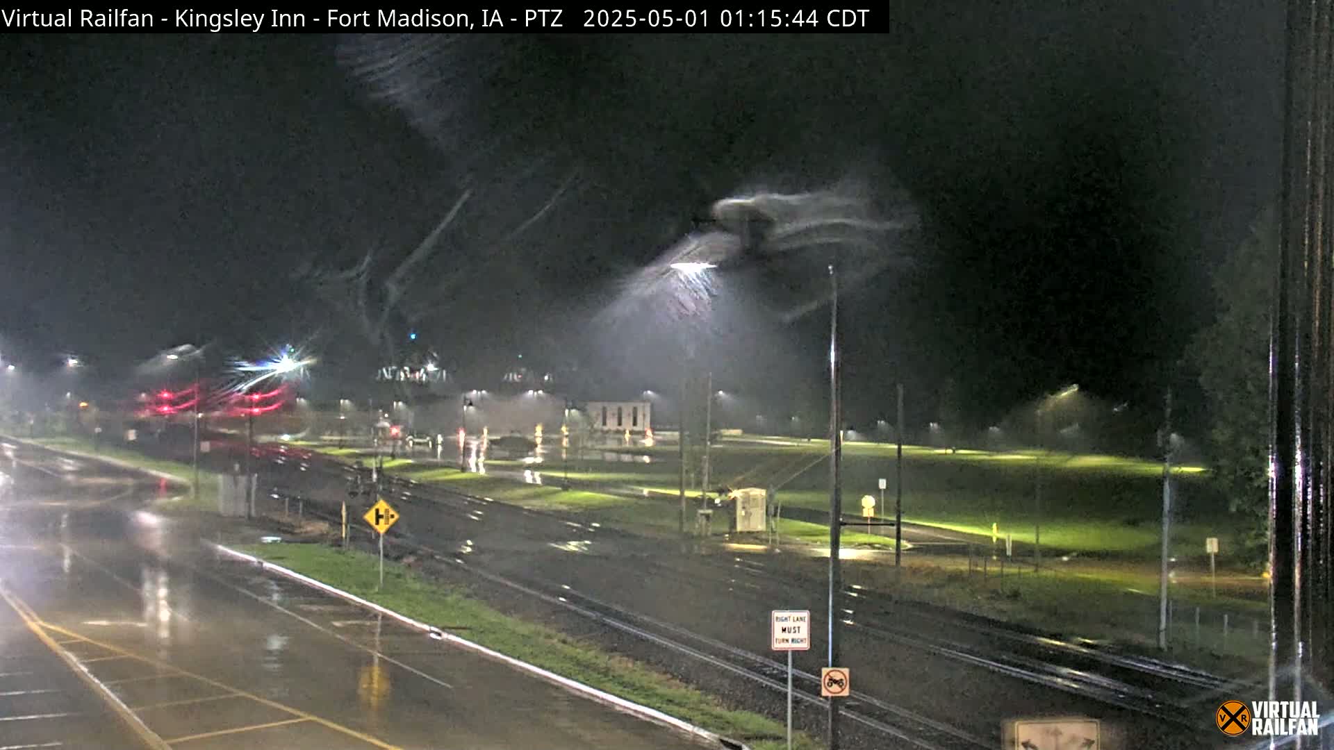 A nighttime view of a rain-soaked intersection with railroad tracks, streetlights, and sparsely-lit buildings under a dark, stormy sky.