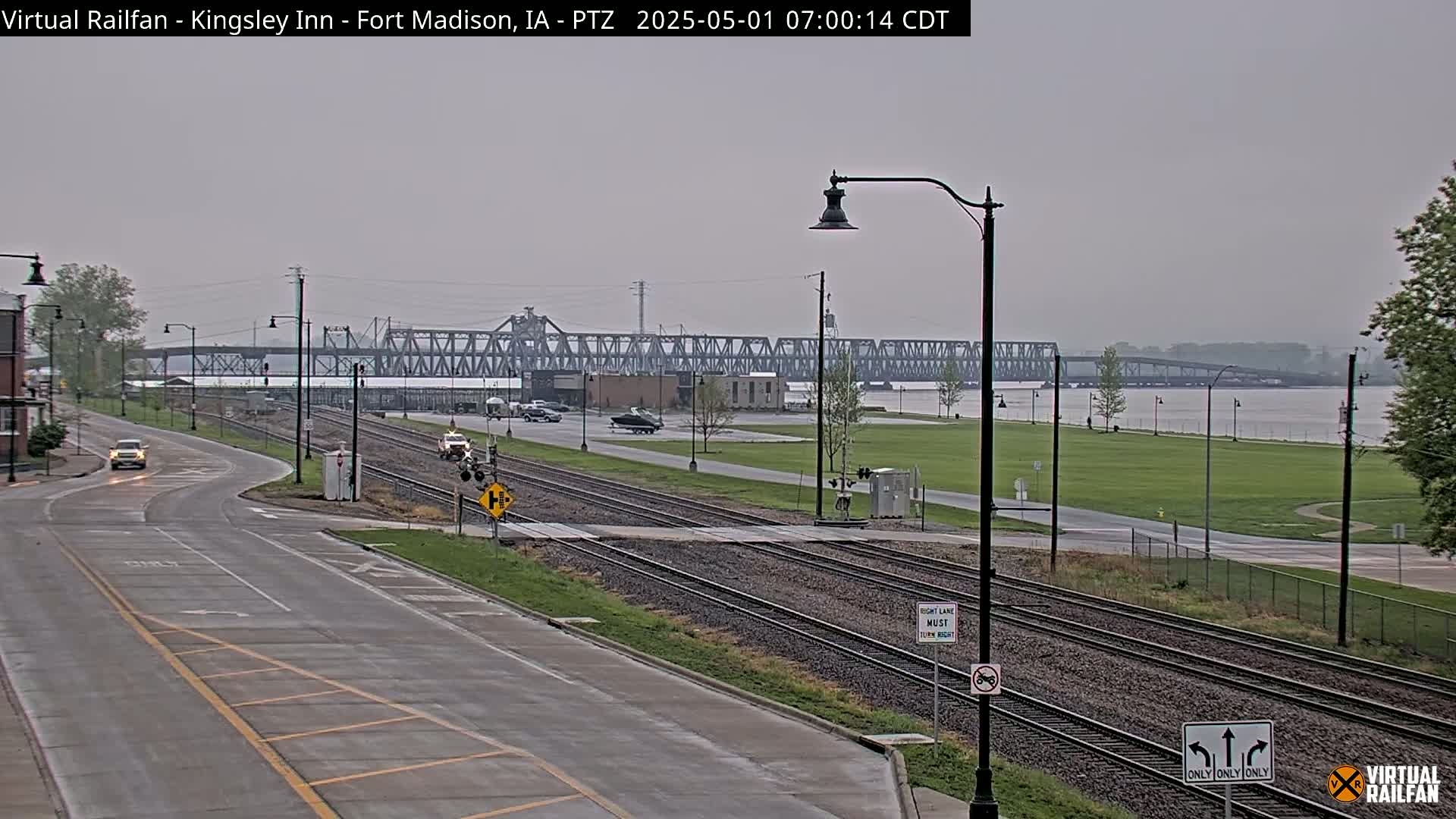 A mostly cloudy day reveals a wide road intersecting train tracks that lead to a large metal bridge spanning a river, with a few cars visible on the road and bridge.