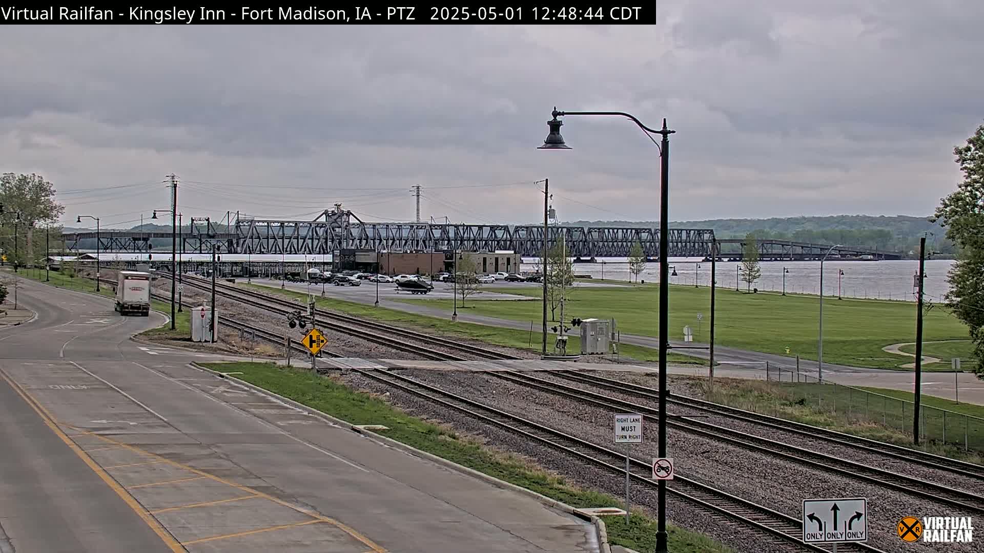 A large metal bridge spans a wide river under an overcast sky, with train tracks and a road intersecting in the foreground.