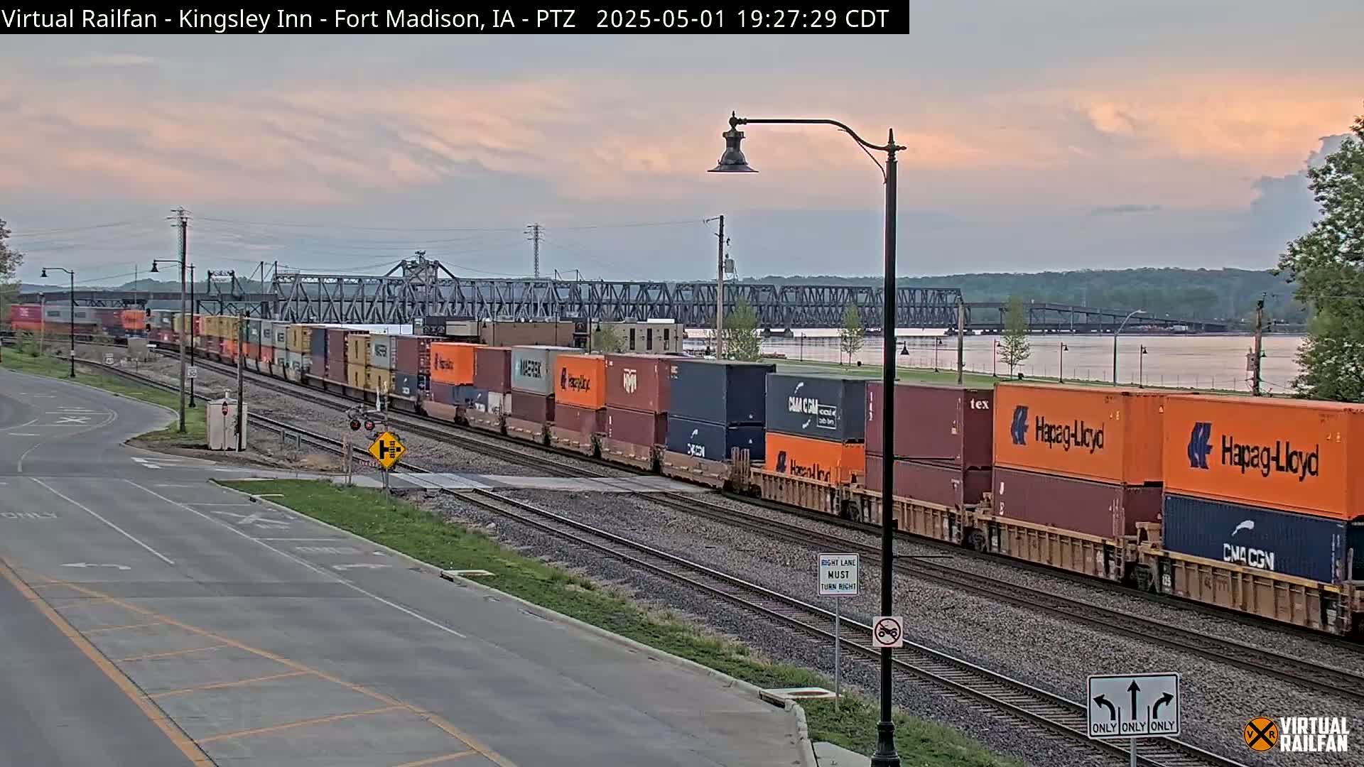 A long freight train carrying many shipping containers passes by a road under a twilight sky near a river and bridge.