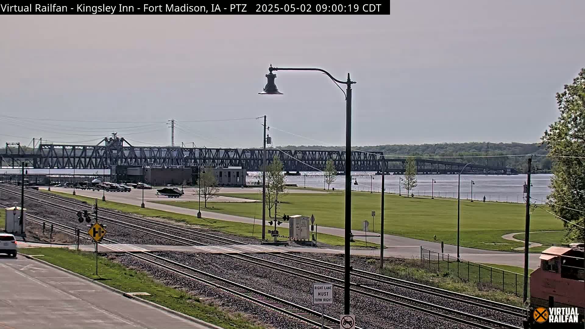 A view of railroad tracks intersecting a road, with a large metal bridge spanning a river in the background under a partly cloudy sky.