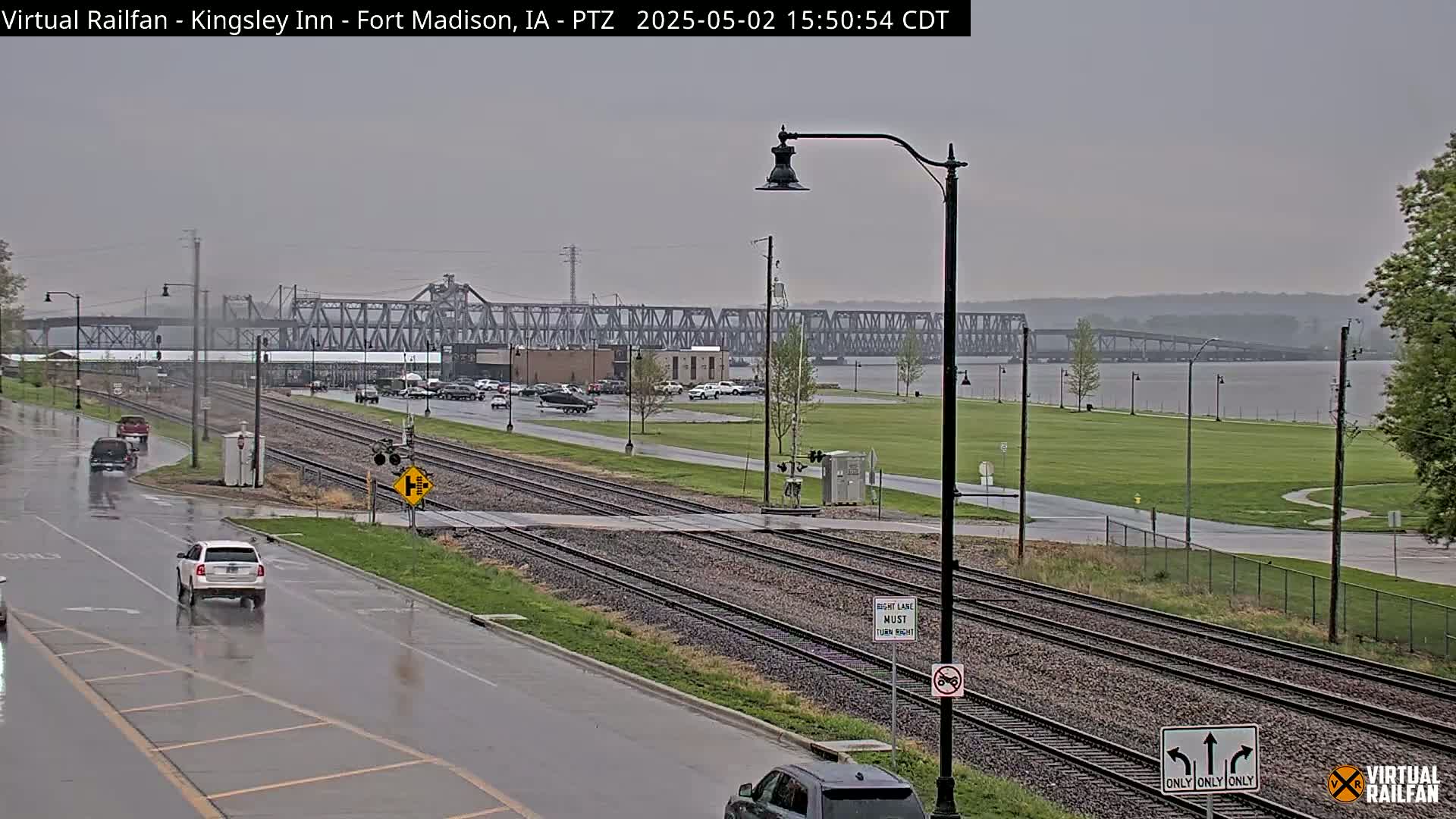 A wet, overcast day is shown, with cars driving on a city street that crosses railroad tracks near a river and a large metal bridge.