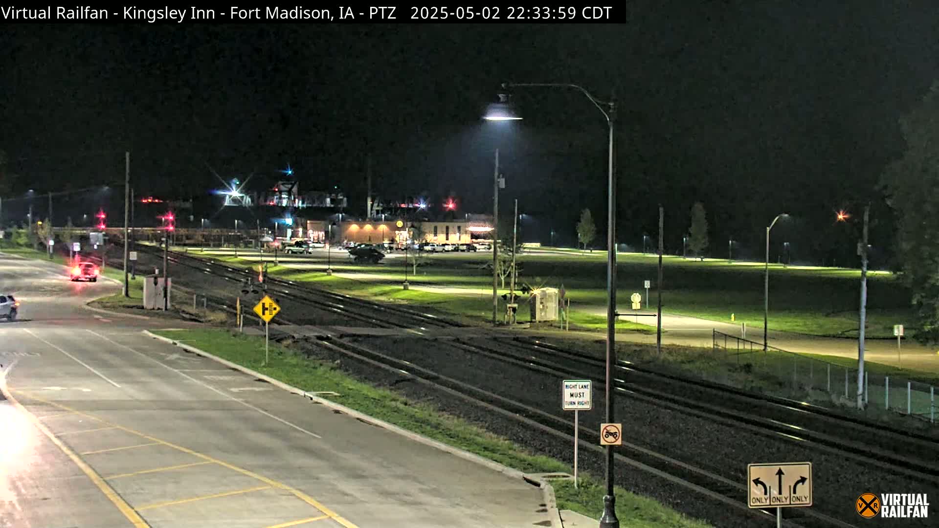 A nighttime view of a road and railroad crossing, with a few cars visible, illuminated by streetlights.