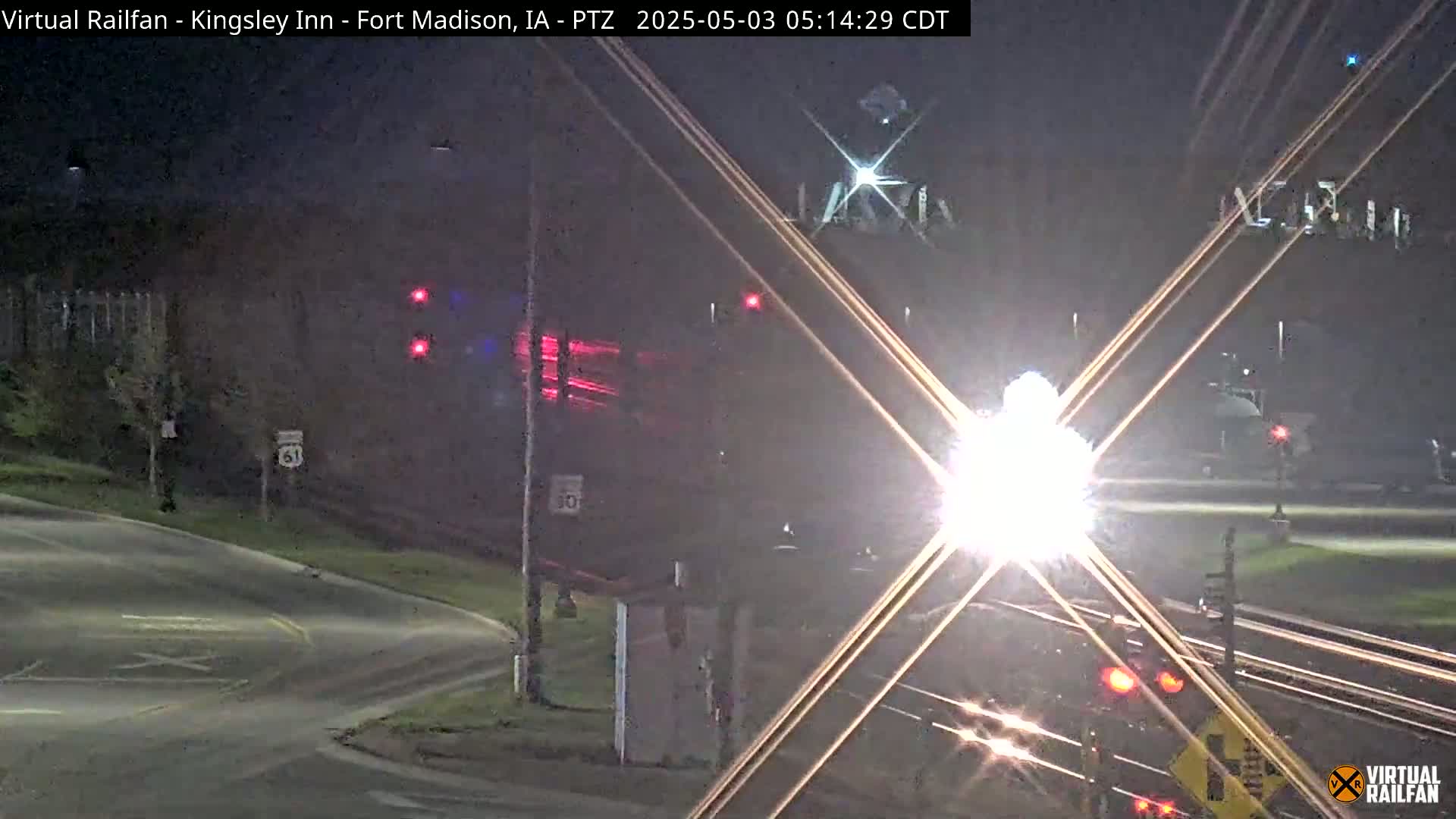 A nighttime view of a train crossing a road intersection, with bright headlights and red traffic signals visible.