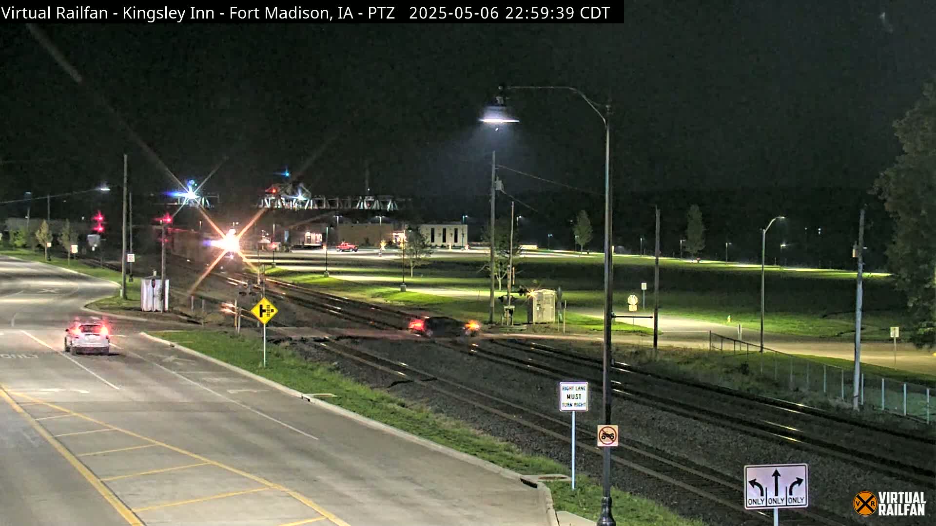 A nighttime scene shows a train passing through a railroad crossing near a road intersection with cars visible, under clear skies.