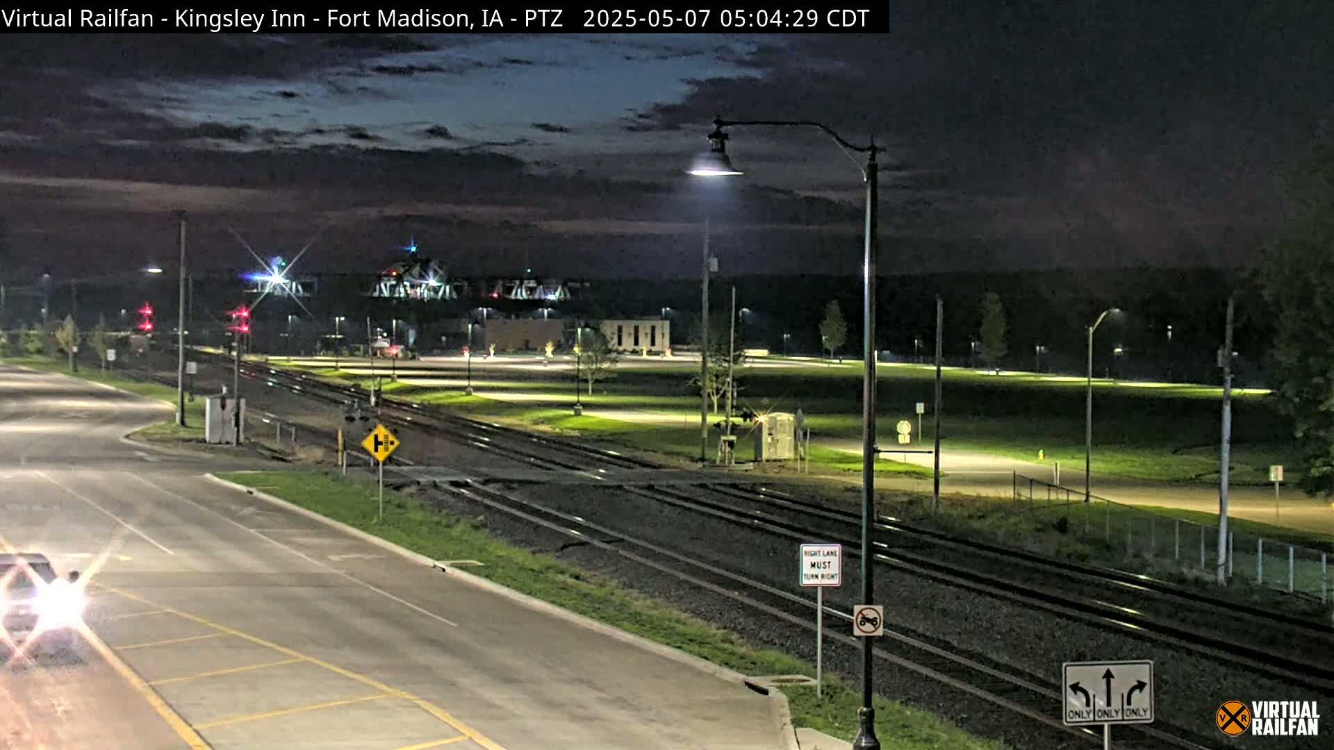 A nighttime view of a road intersecting railroad tracks near a bridge, under a mostly clear sky.
