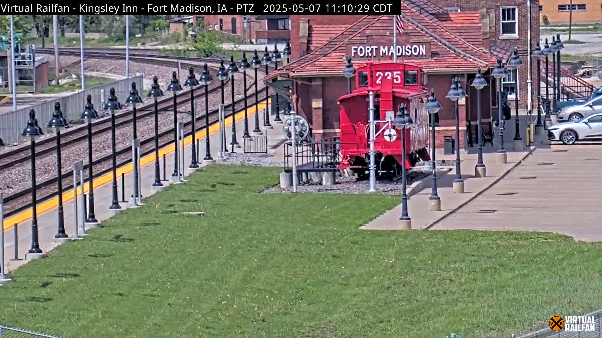 A red caboose sits beside a train station on a sunny day, with numerous lampposts lining a paved walkway next to train tracks.
