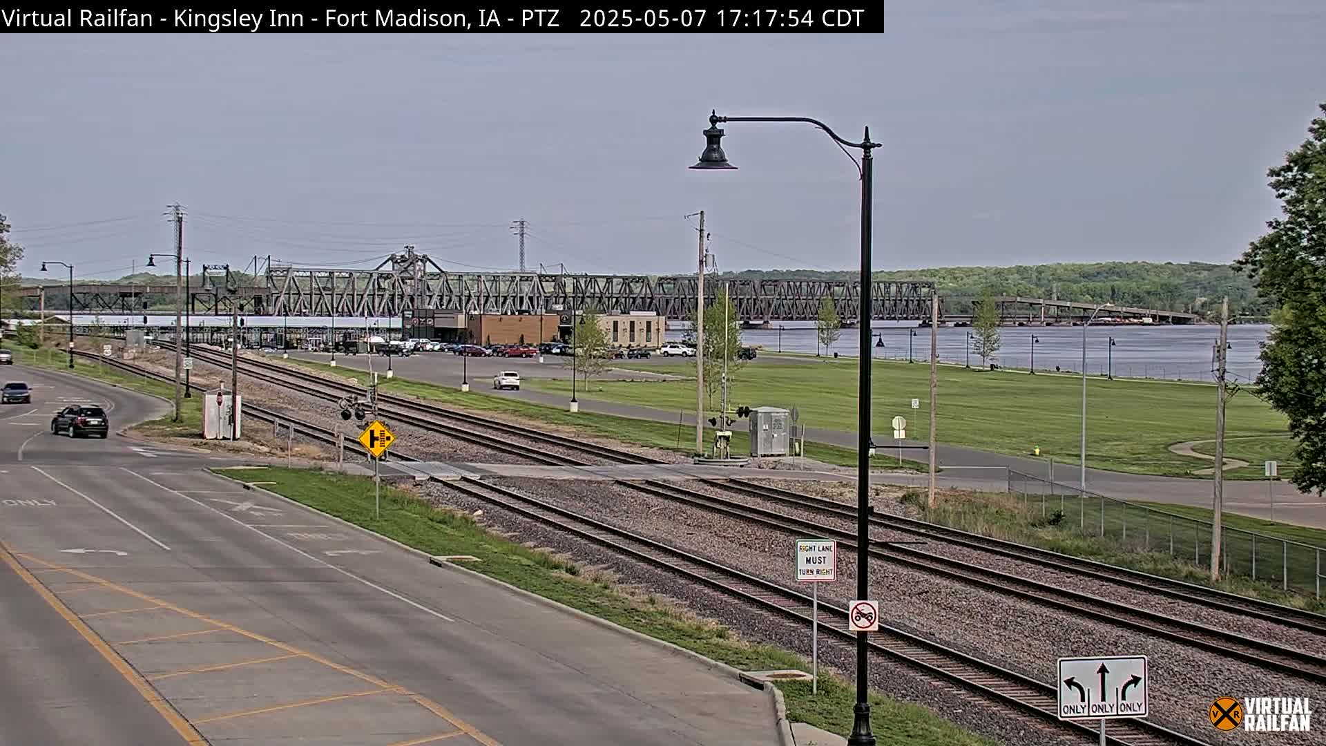 A mostly sunny day shows a view of railroad tracks crossing a road, with a river and a large bridge in the background.
