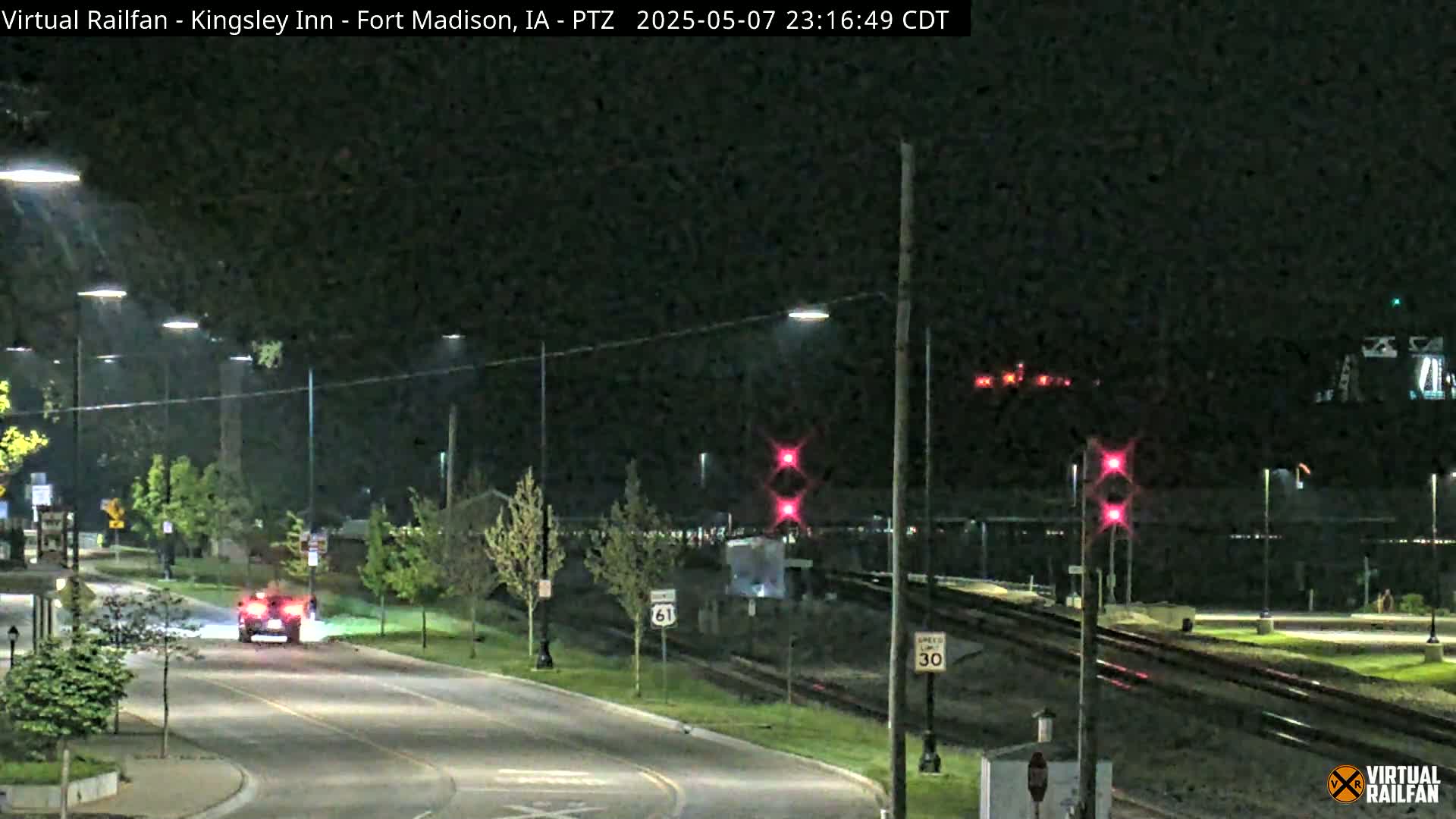 A nighttime view of a street and railroad crossing with a car driving on the street and a train in the distance, under streetlights.