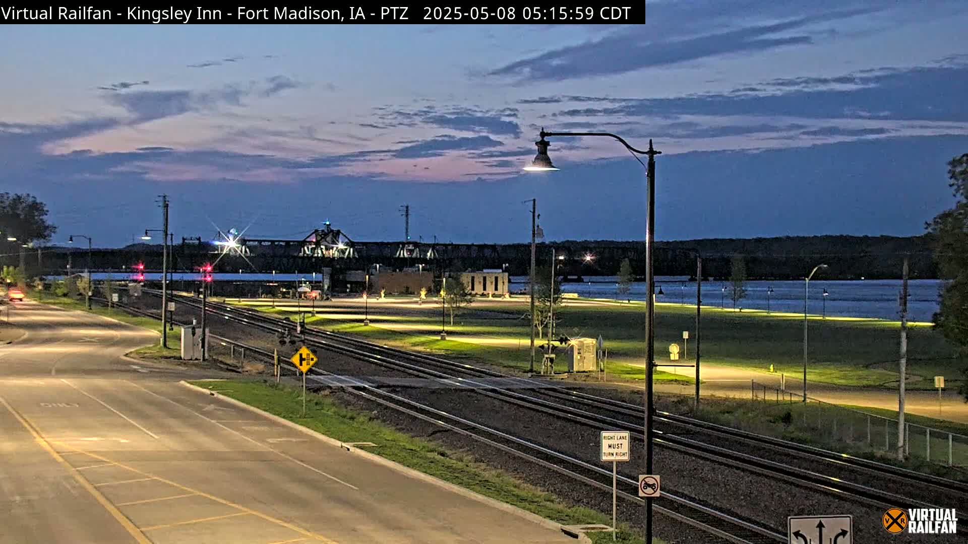 A twilight view shows a roadway intersecting railroad tracks near a river, with a bridge and a building visible in the background under a mostly clear sky.