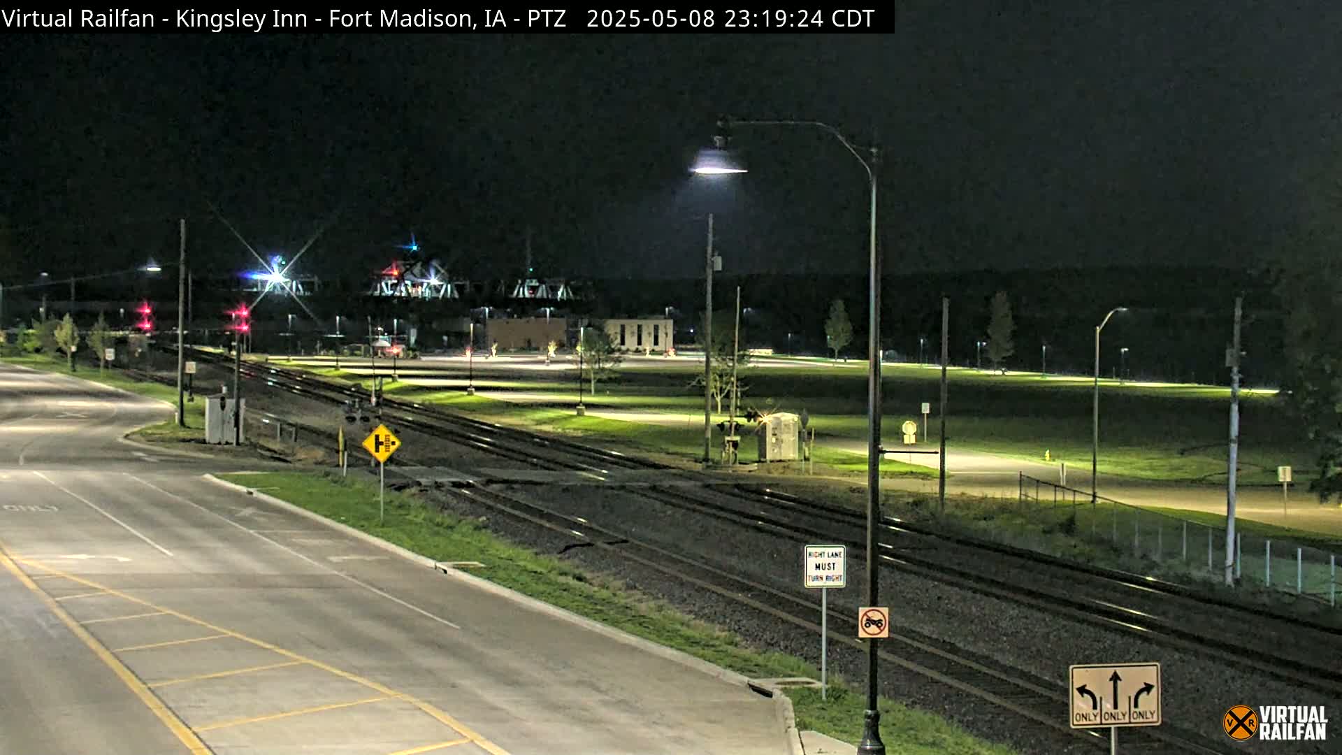 A nighttime view of a road and railroad crossing, illuminated by streetlights, with a grassy area and buildings in the background.