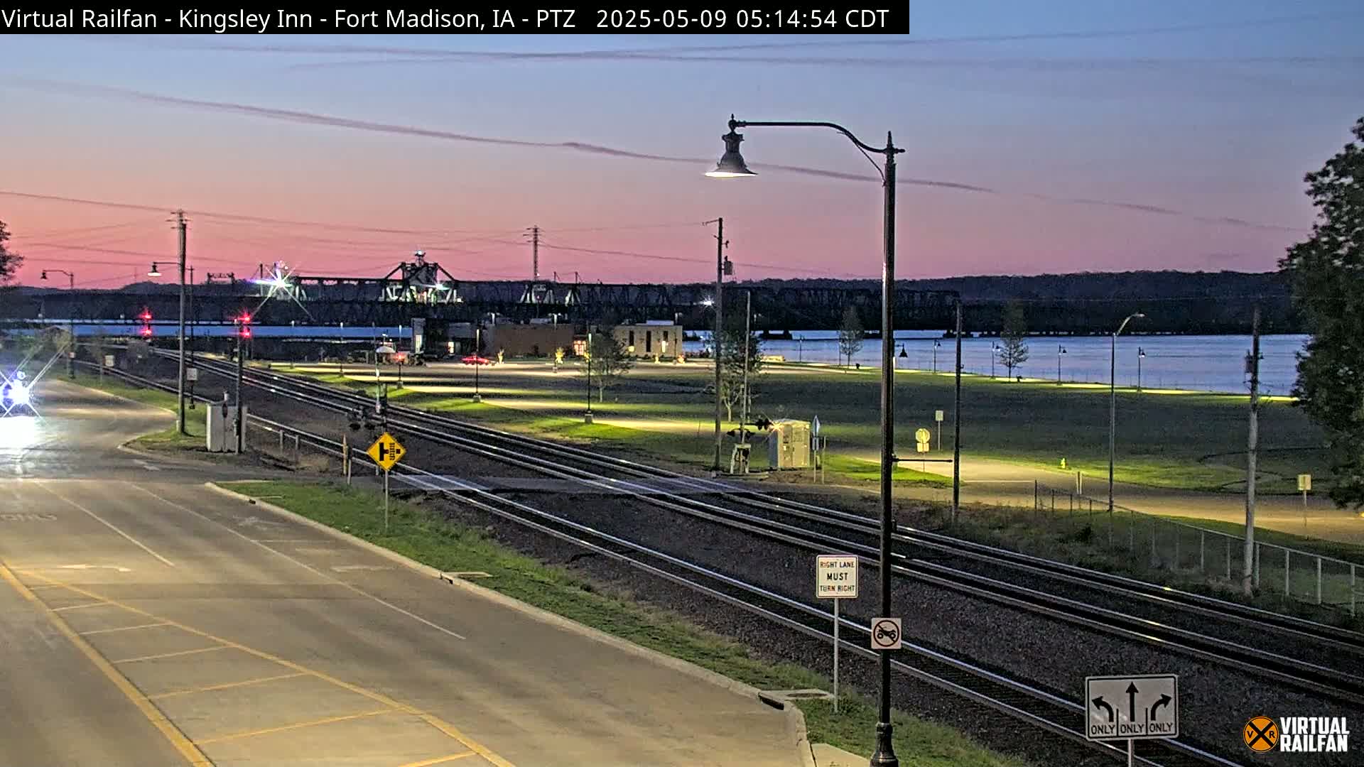 A twilight view shows railroad tracks crossing a road next to a river, with a bridge in the background under a pinkish-purple sky.