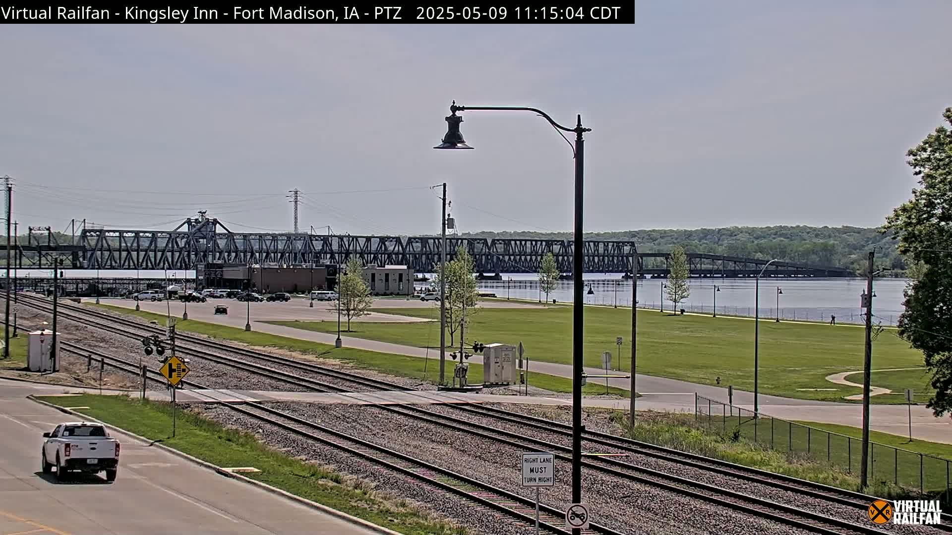 A view of a river, a large metal bridge, railroad tracks, and a roadway under a partly sunny sky.