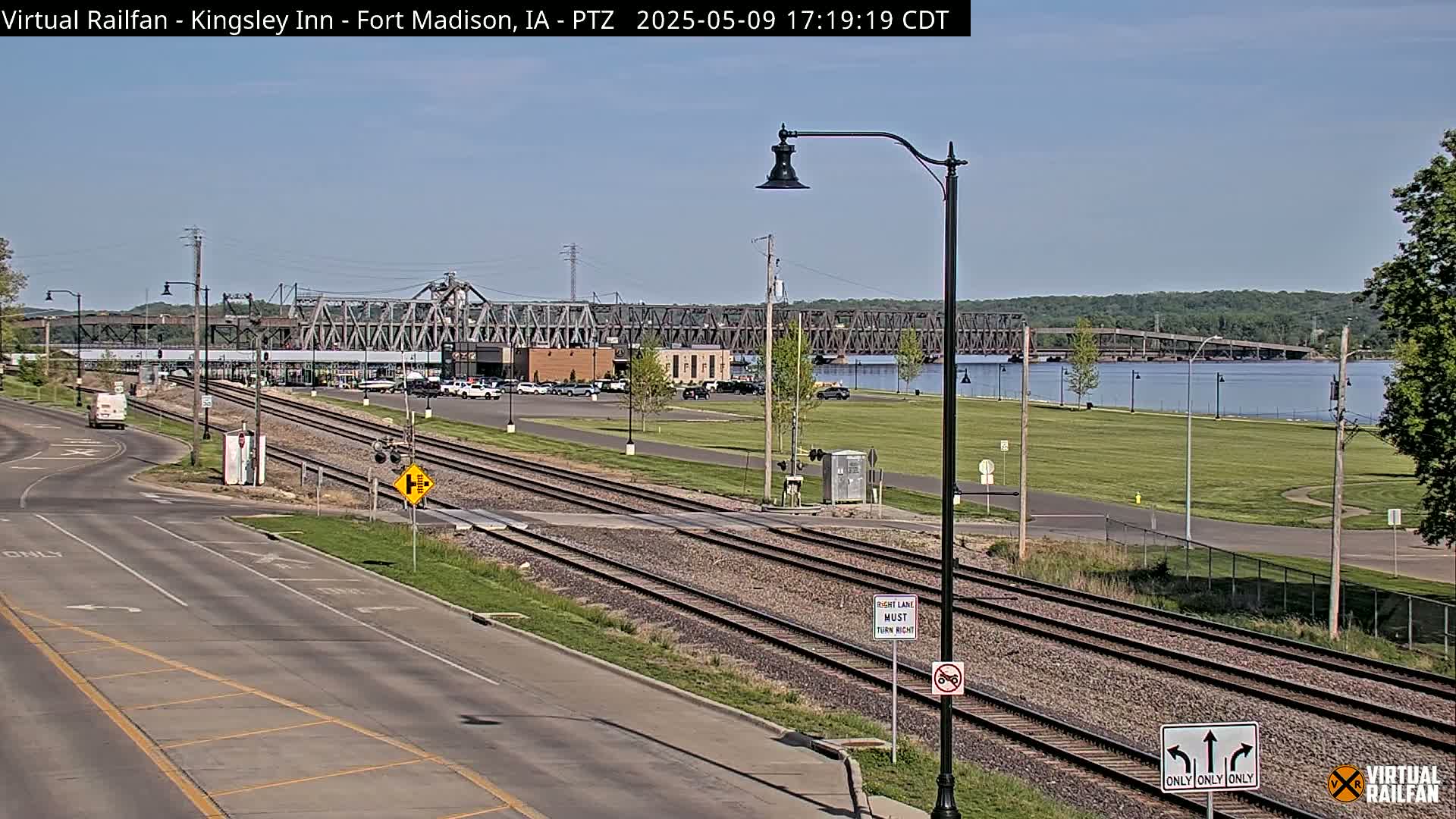 A view of railroad tracks intersecting a road, with a metal bridge and calm river in the background under a clear sky.