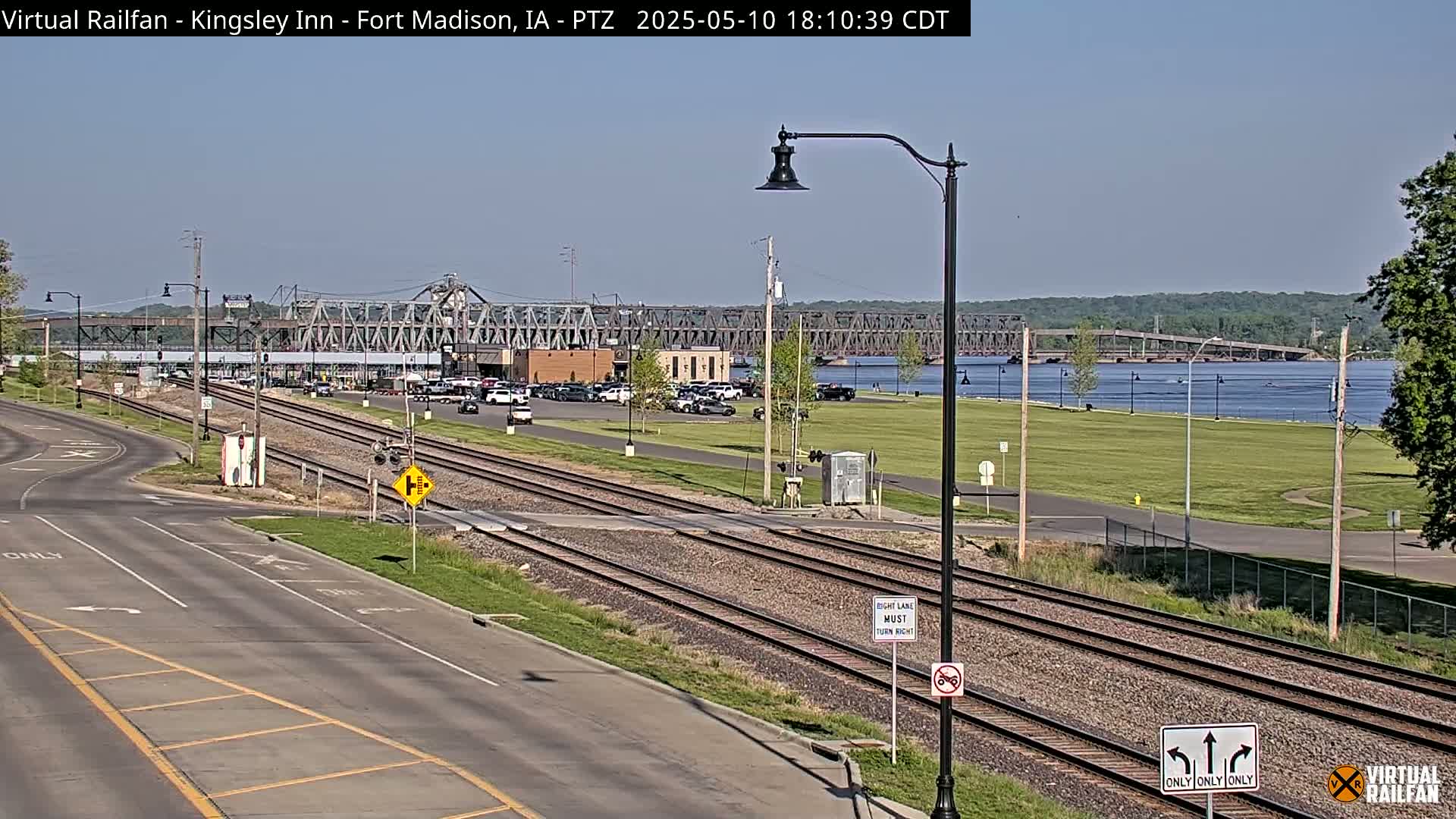 A long, mostly empty roadway with train tracks running parallel, leading to a large metal bridge spanning a wide river on a sunny day.