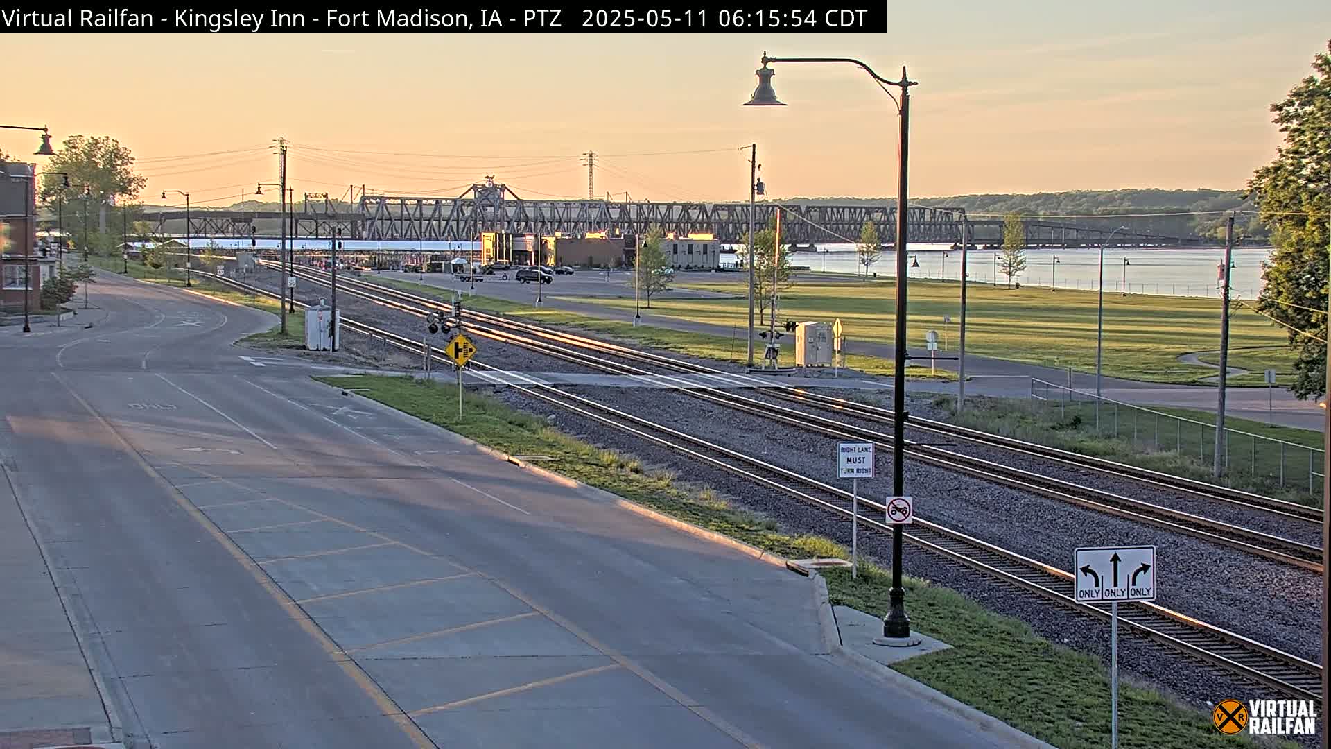 A sunrise illuminates a road intersecting with train tracks near a river and bridge.