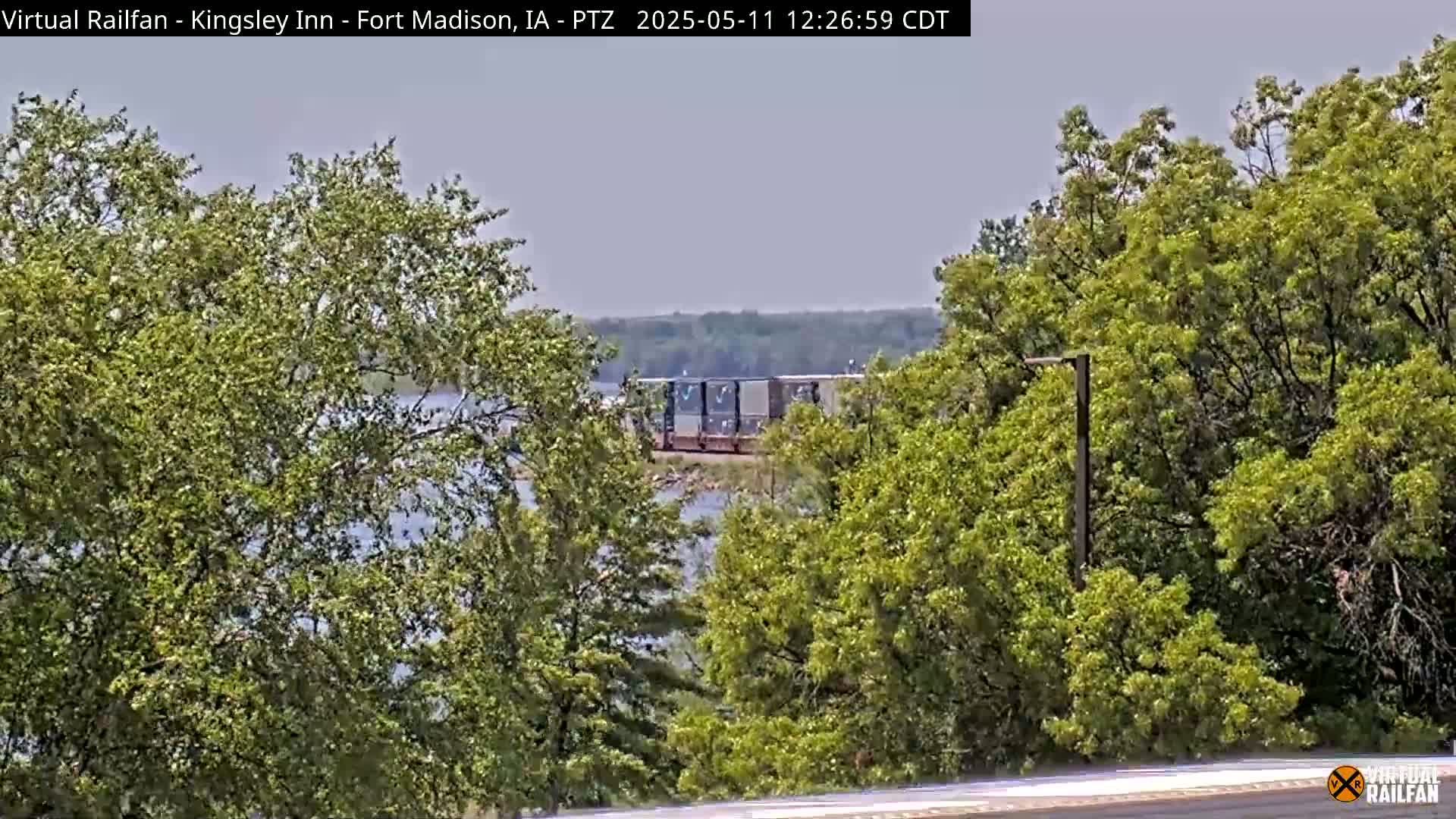 A freight train is visible beyond a body of water and through leafy green trees under a hazy sky.