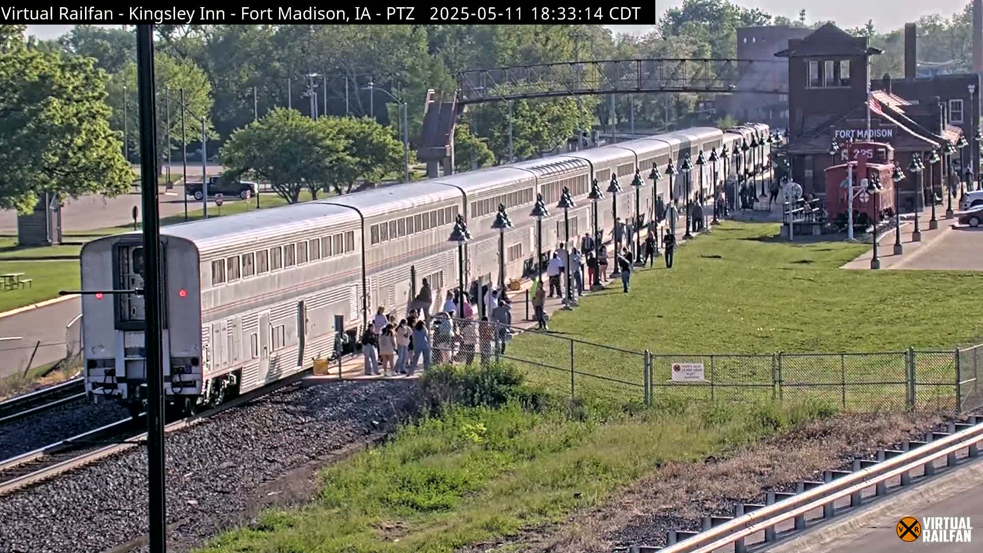 A long silver passenger train is at a station platform where many people are standing, on a sunny day.