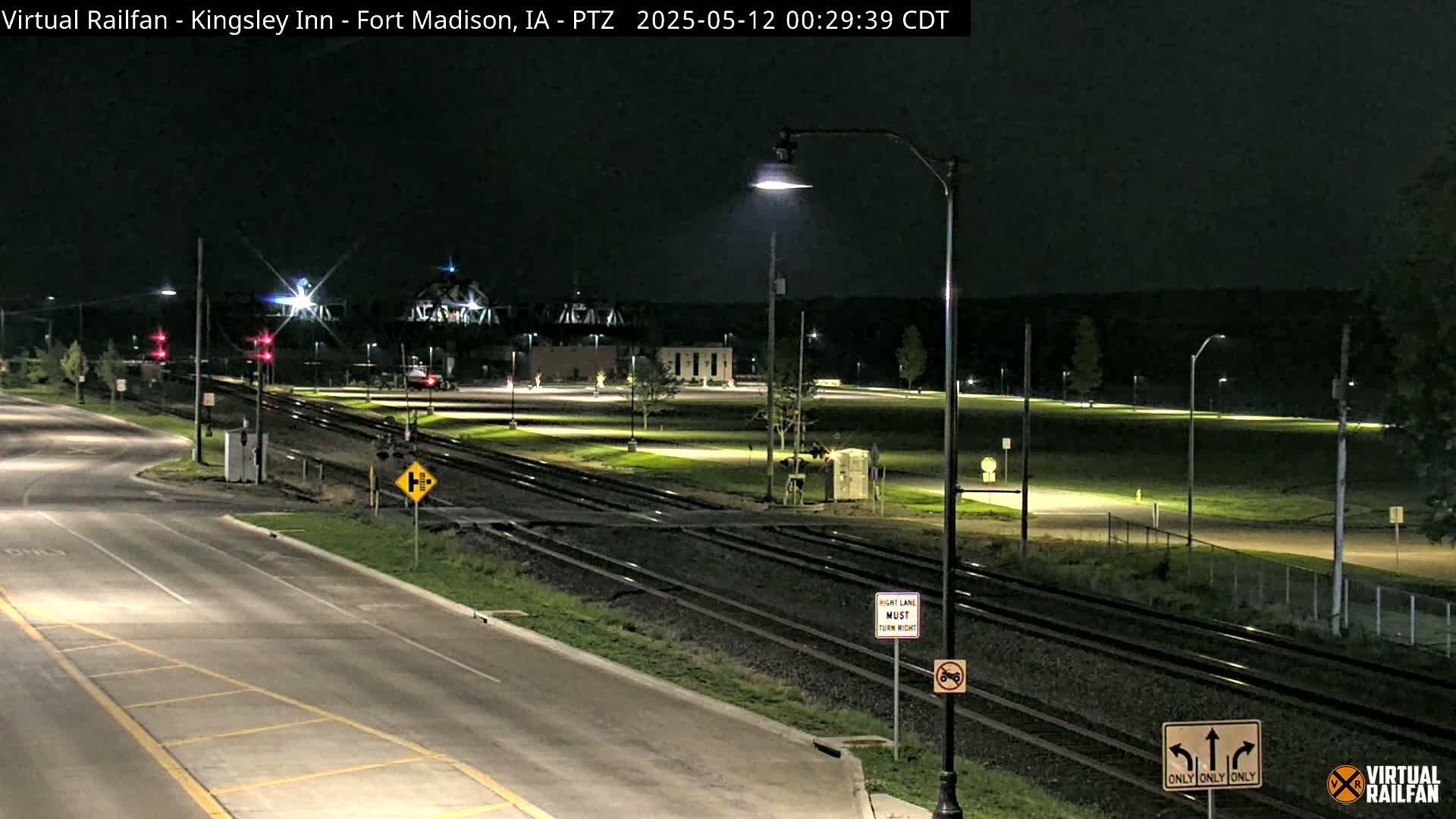 A nighttime view of a road intersection with railroad tracks, featuring streetlights illuminating a mostly empty parking lot and surrounding grassy areas.