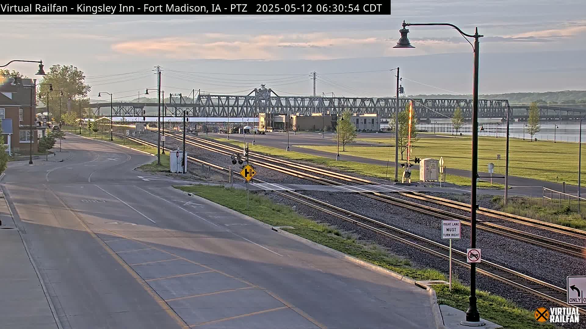 A mostly clear dawn illuminates a roadway intersecting railroad tracks that lead to a metal truss bridge over a river.