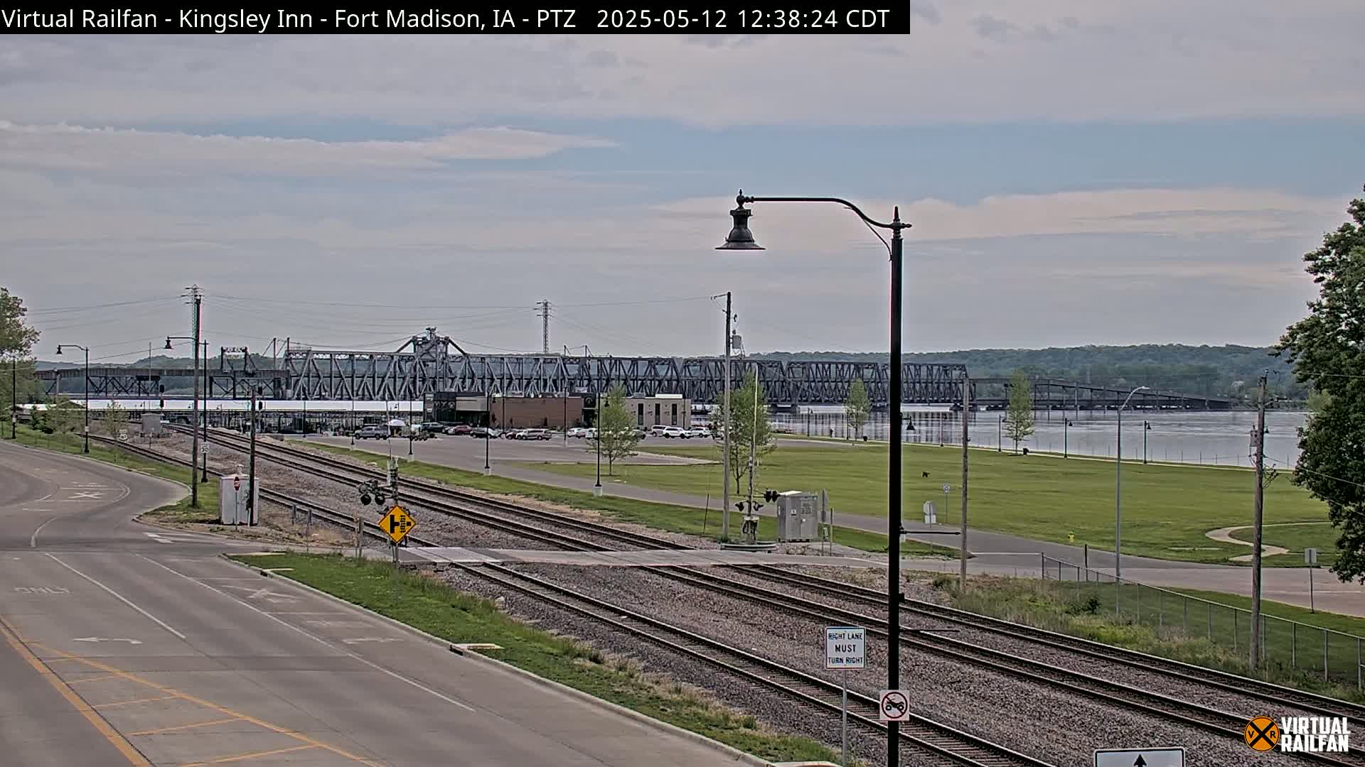 A view of a river with a metal bridge in the background, train tracks and a road in the foreground under a partly cloudy sky.