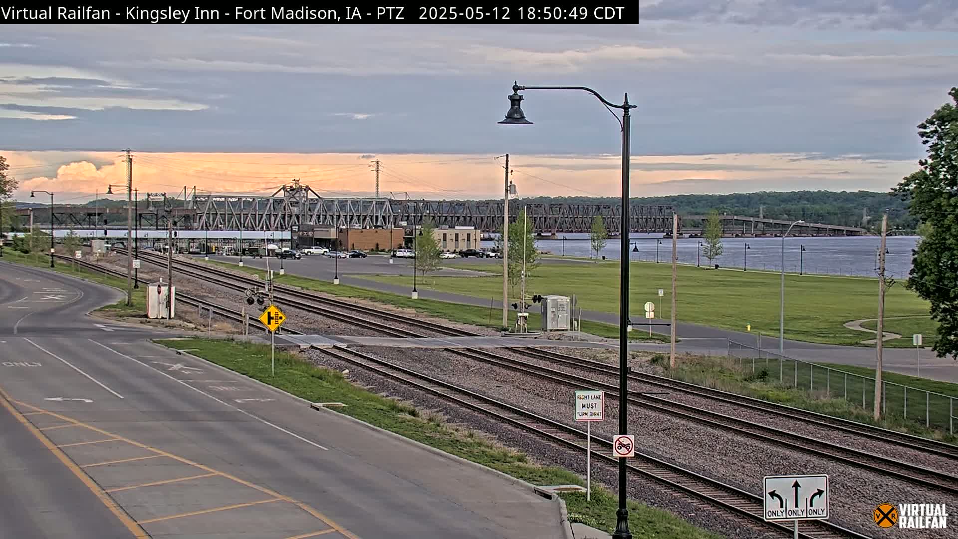 A view of a railroad crossing near a river at sunset, with a large bridge in the distance.
