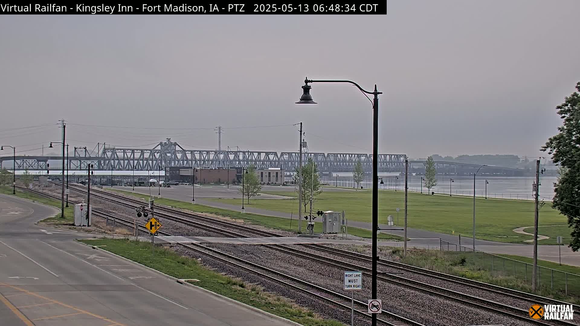 A hazy view of a railroad crossing with train tracks leading toward a large metal bridge over a river, under an overcast sky.