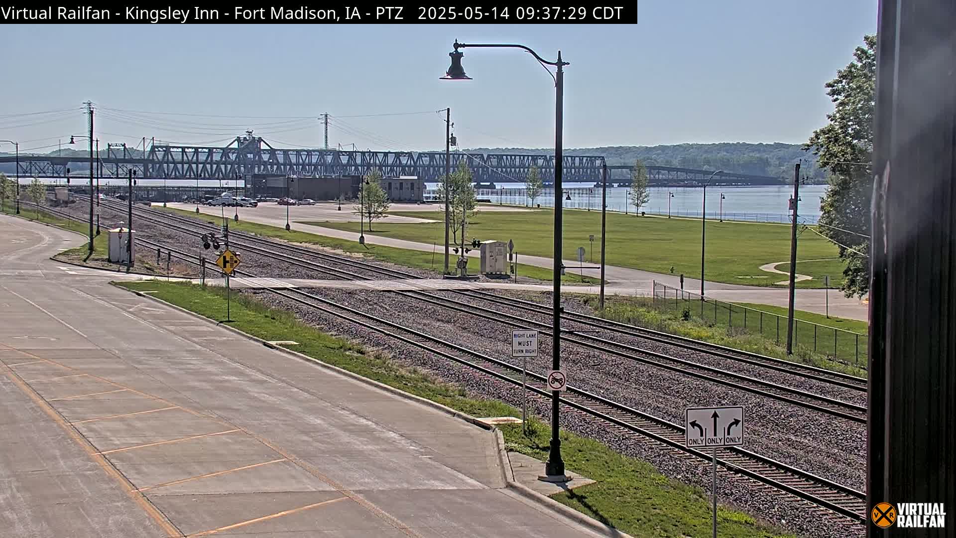 A sunny view of railroad tracks intersecting a road, with a large metal bridge spanning a river in the background.