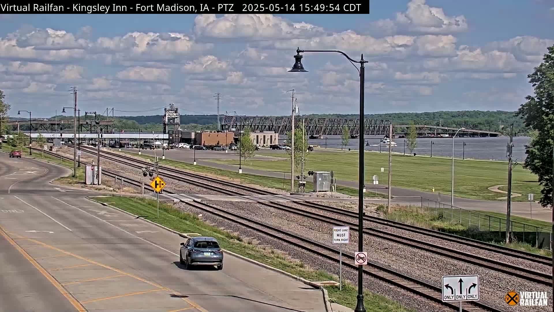 A partly cloudy day shows a view of a river, a bridge, railroad tracks, and a road with a vehicle driving on it.