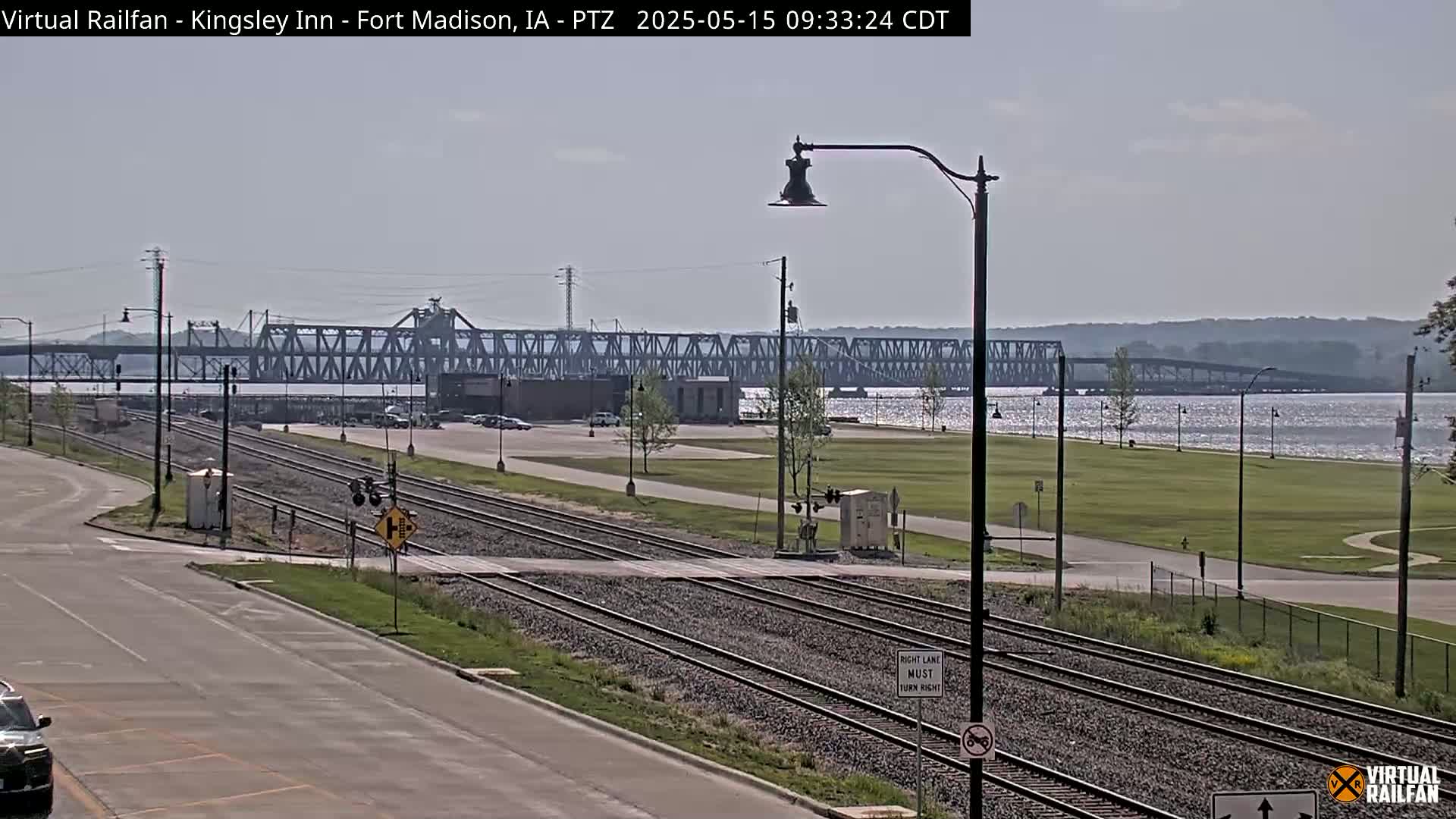 A view of railroad tracks crossing a road, with a large metal bridge spanning a body of water in the background on a sunny day.