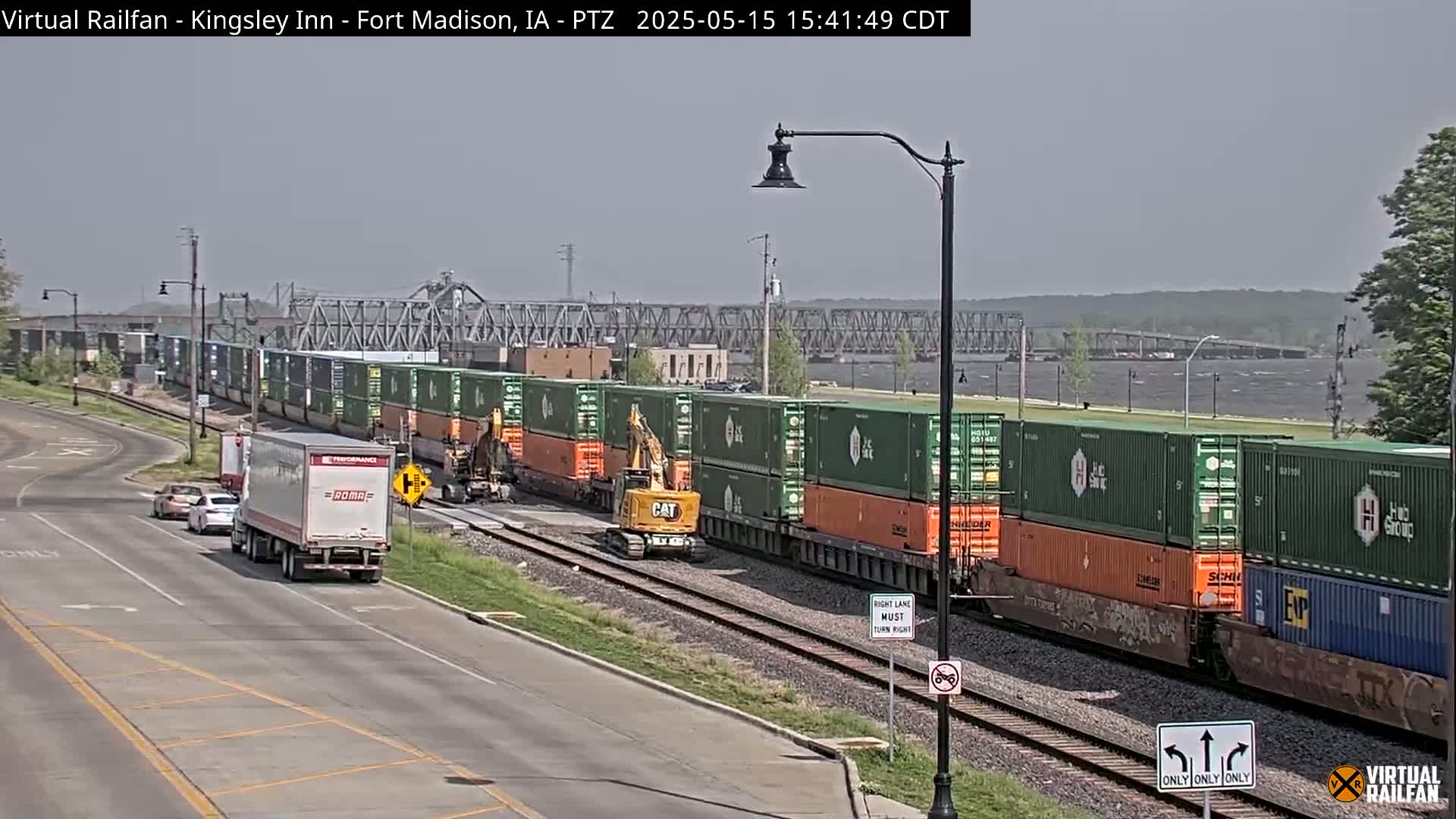 A long freight train carrying numerous shipping containers passes by a road with several cars and trucks, under a mostly clear sky next to a river and bridge.