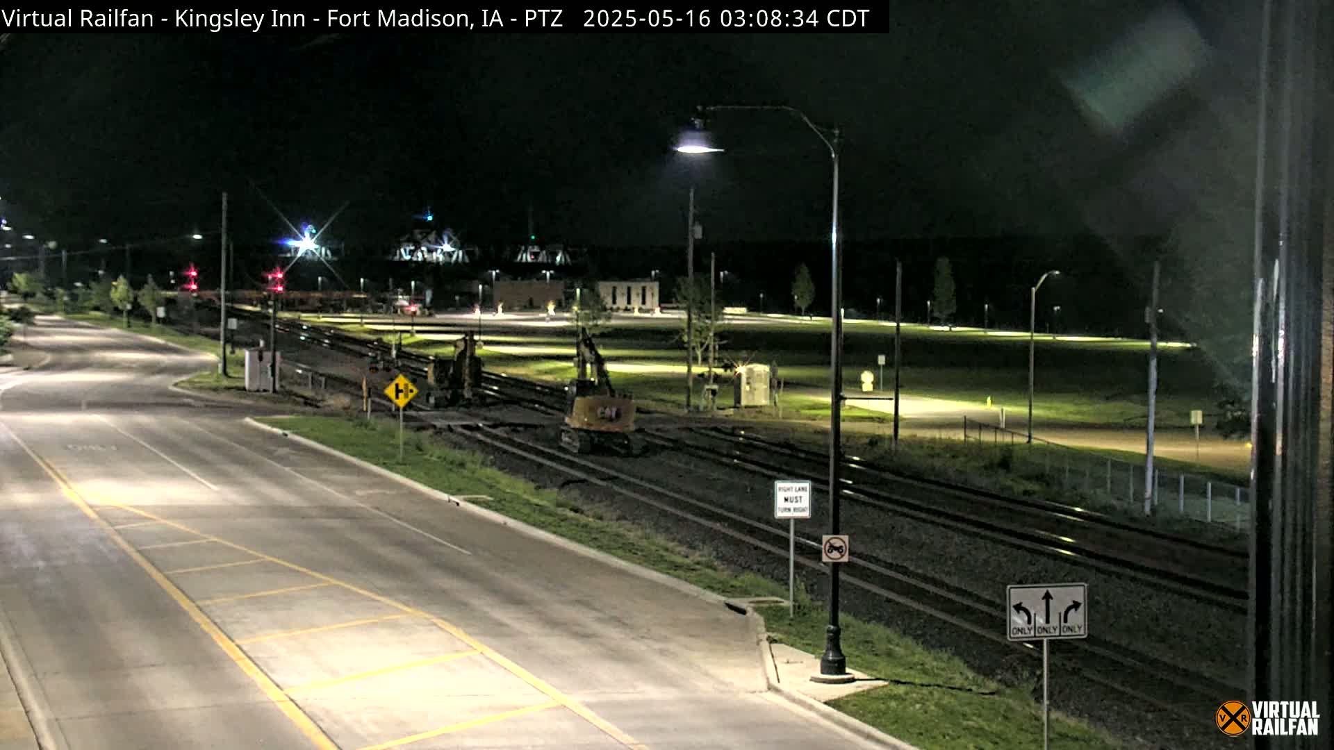 At night, under dark skies, construction equipment sits on railroad tracks next to a road, with lights illuminating the scene.