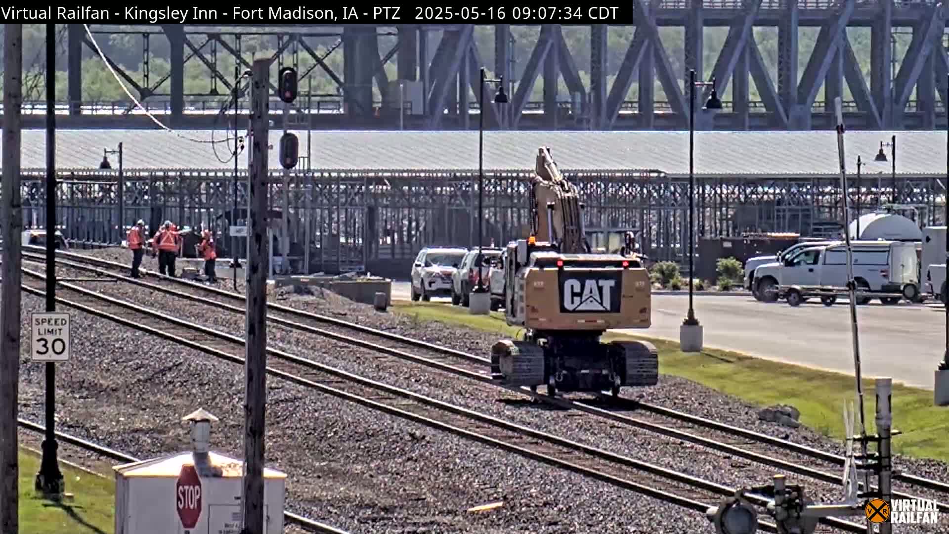 A yellow Caterpillar excavator moves along railroad tracks near a bridge on a sunny day, with several workers and vehicles visible in the background.