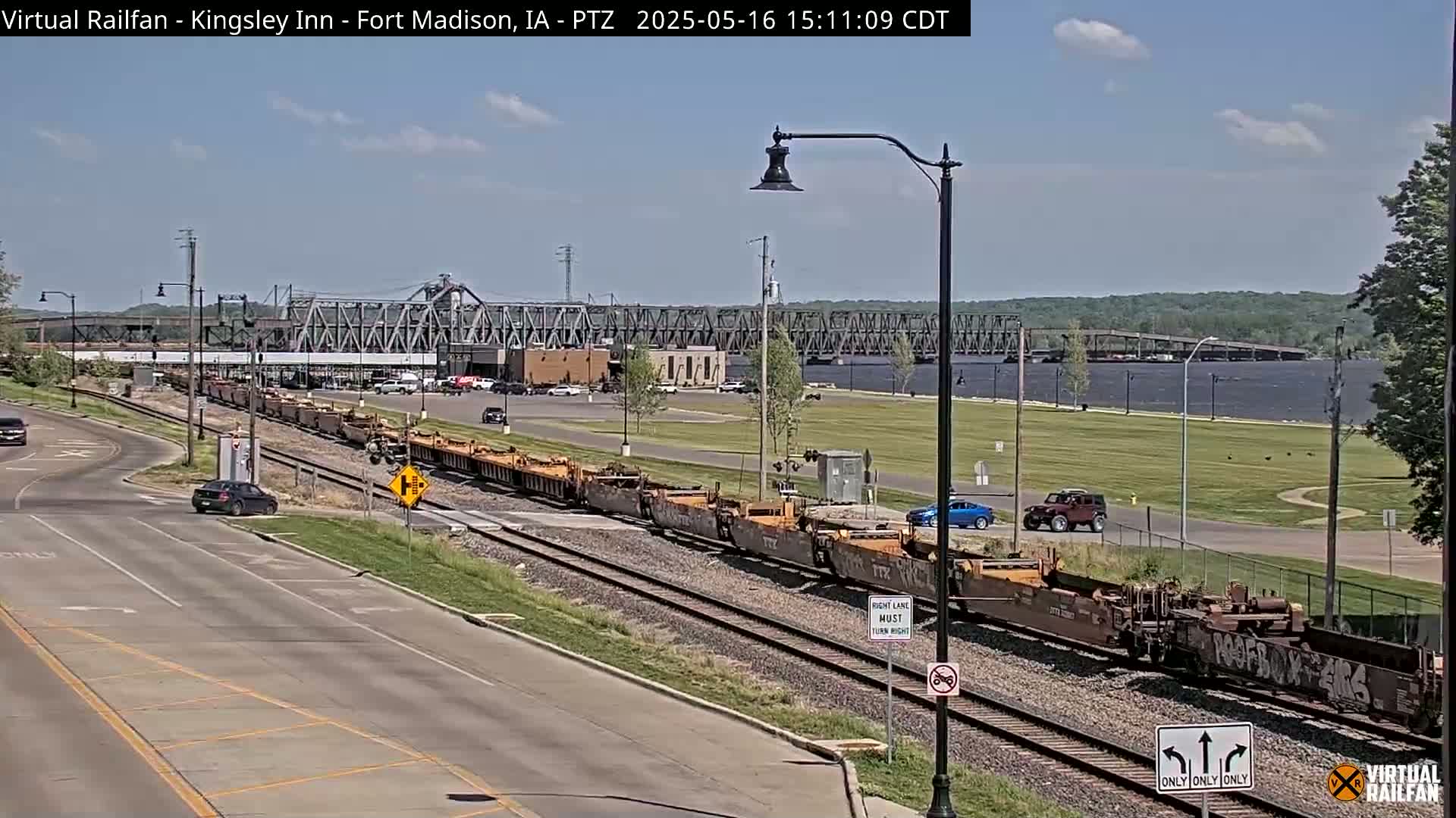 A long freight train is on a set of tracks near a river under a mostly sunny sky, with several cars visible on a road nearby and a bridge in the background.