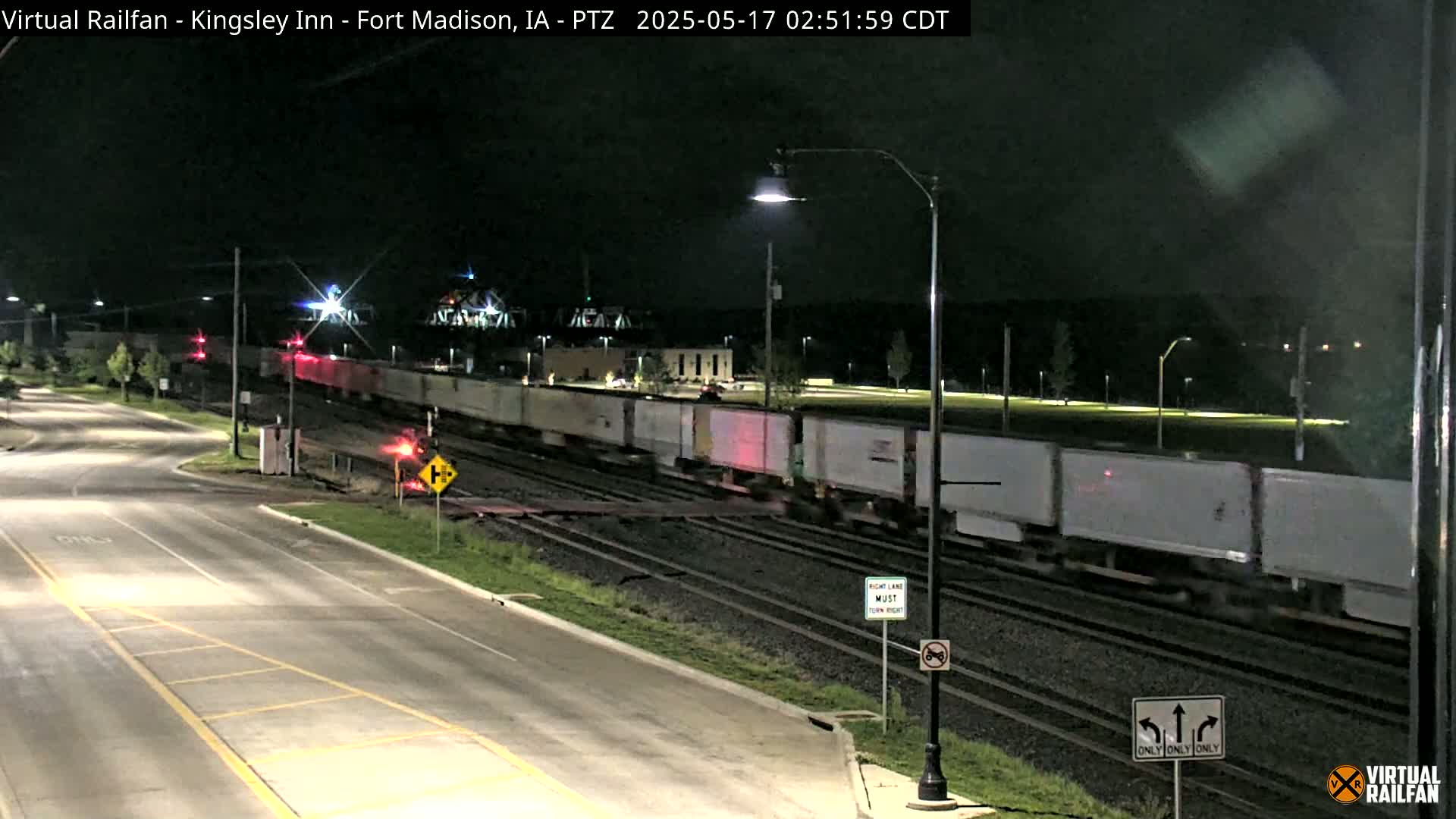 A long freight train moves through a railroad crossing at night under streetlights.