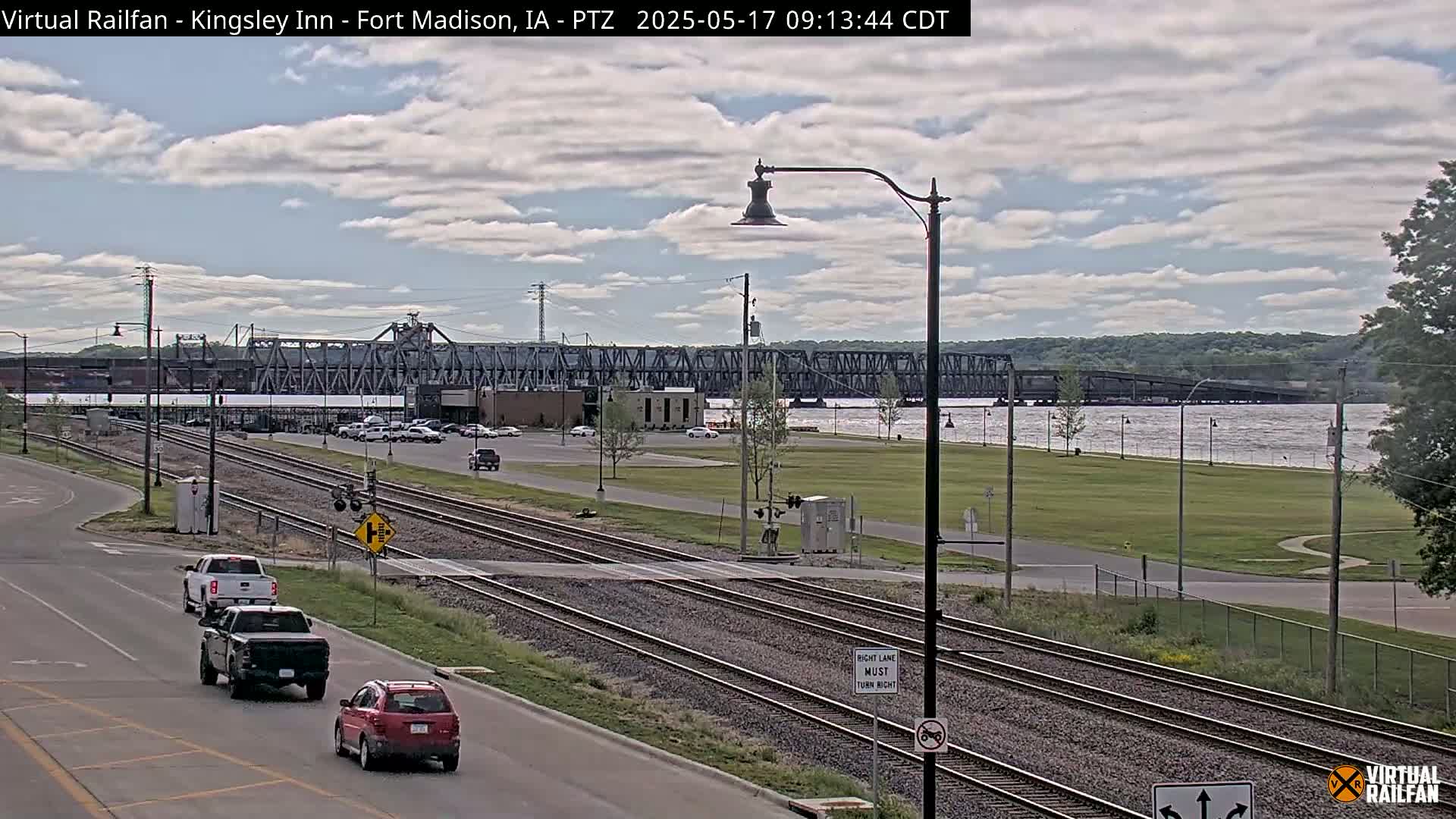 A partly cloudy day shows a view of a river with a large metal bridge, railroad tracks, and several vehicles on a road near a railway crossing.